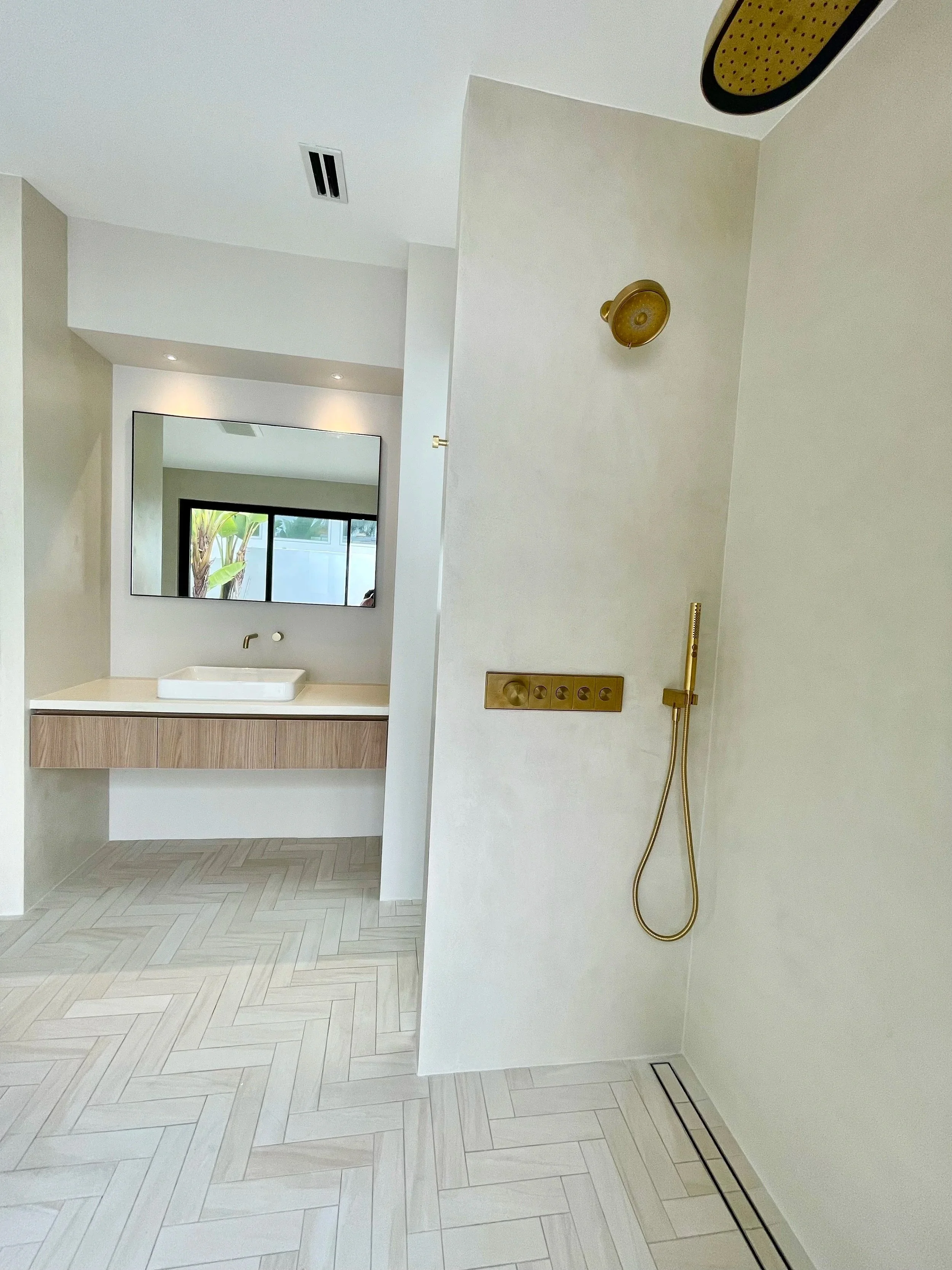 Modern Tadalekt plaster  bathroom with a walk-in shower featuring gold fixtures, a wooden vanity with a white sink, a large mirror, and light-colored herringbone tile flooring.