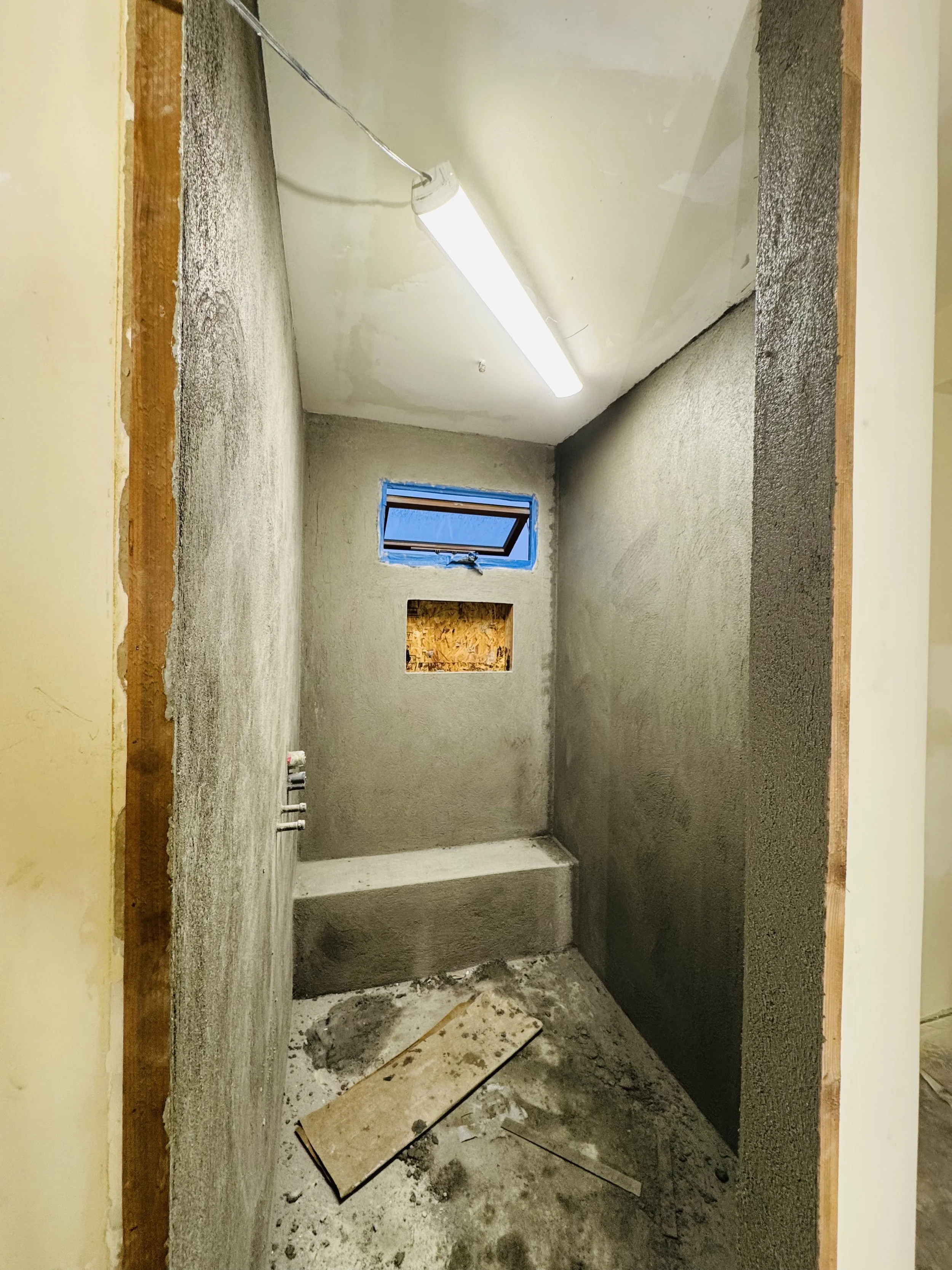 A small, unfinished room under construction with cement walls, a small rectangular window, and exposed wiring. The ceiling has a fluorescent light fixture, and the floor has construction debris.