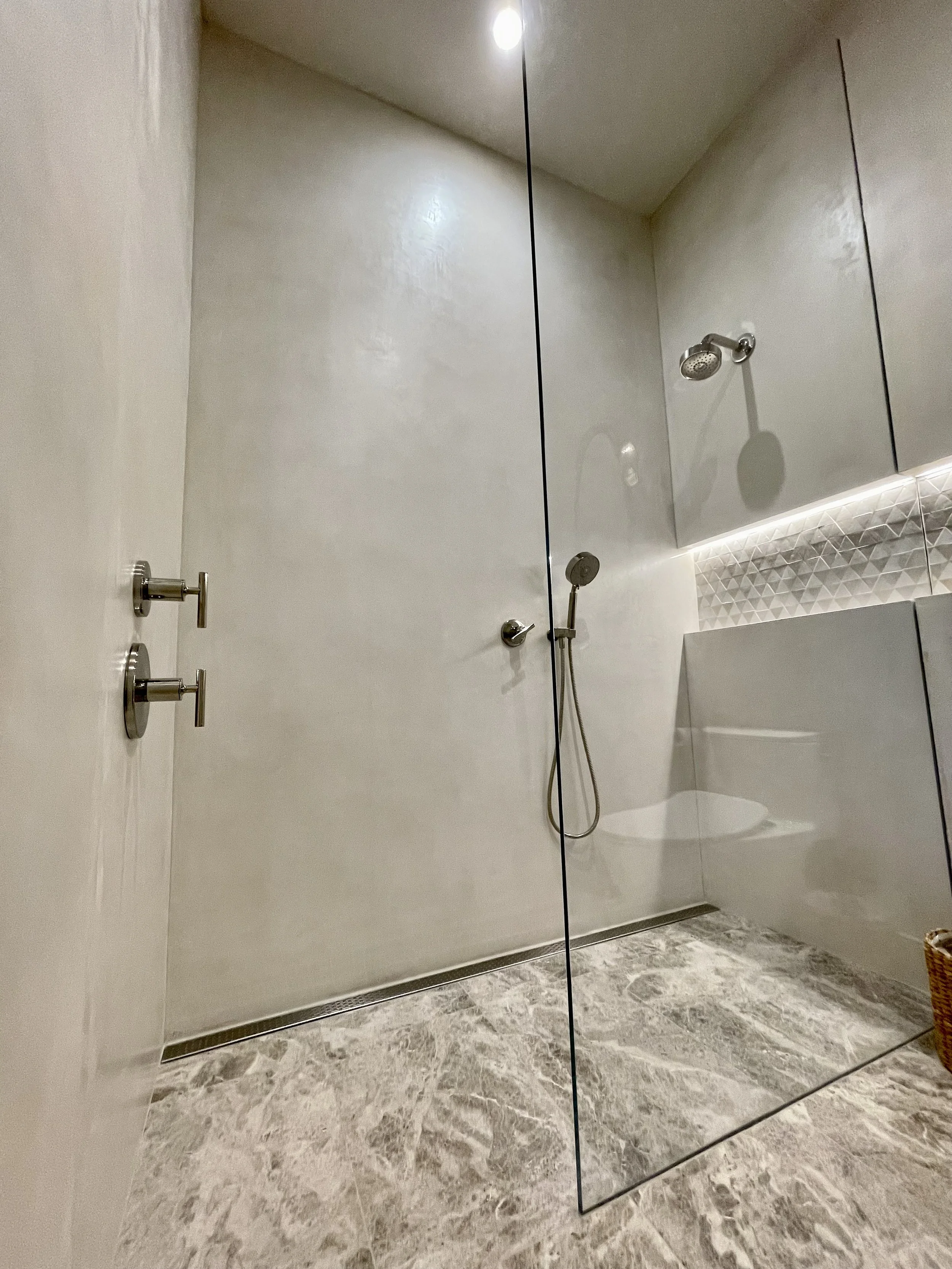 Tadelakt plaster walk-in shower with a glass partition, silver fixtures, a handheld showerhead, and marble flooring in a beige and white color scheme.