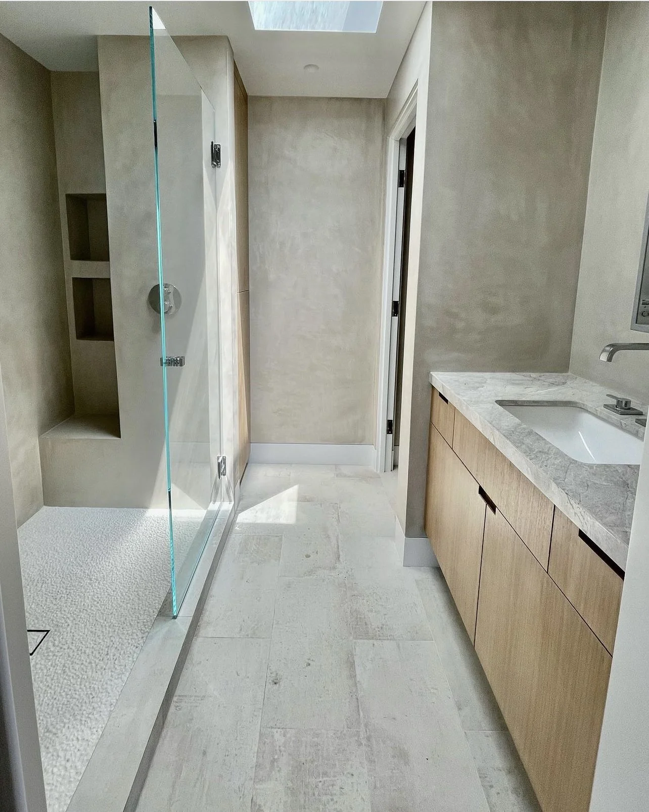 Modern bathroom with a walk-in shower, built-in shelves, a skylight, and a wooden vanity with a marble countertop.