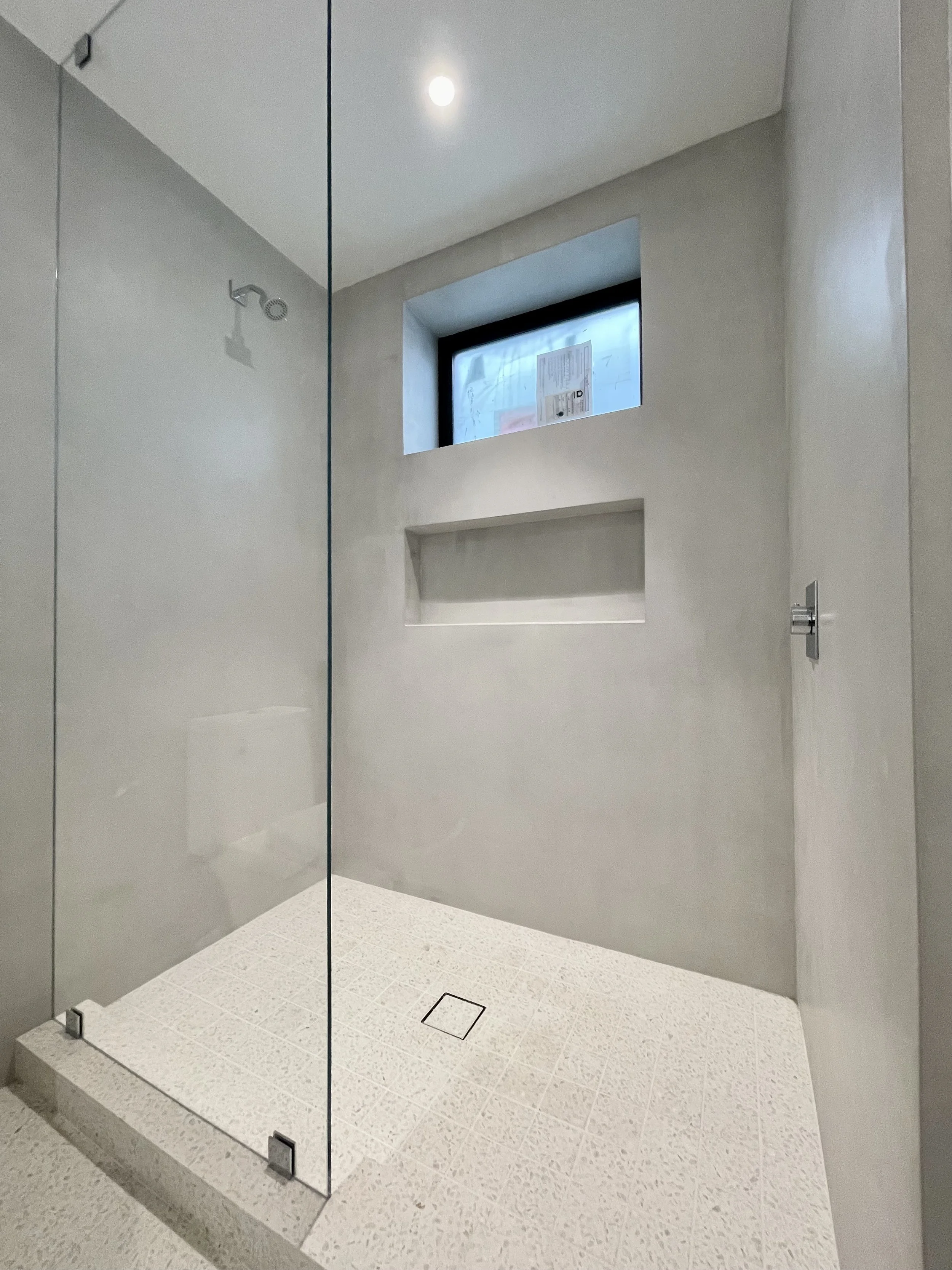 Modern tadelakt plaster walk-in shower with a glass door, a small window, a wall-mounted showerhead, and a built-in shelf, in a minimalist bathroom.