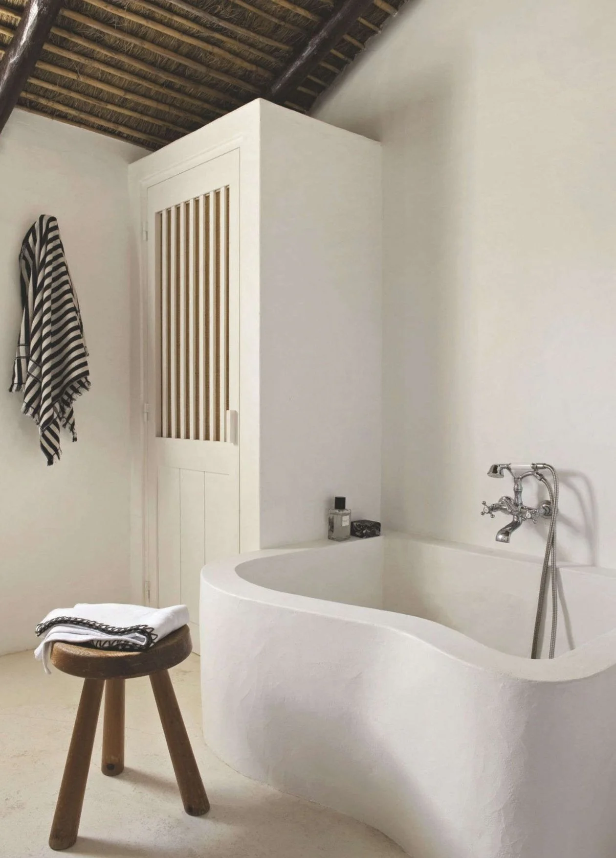 A minimalist bathroom with a curved white bathtub, a wooden stool with a white towel, a striped towel hanging on the wall, and a small white cabinet with wooden slats on the door. The room has white walls, light-colored flooring, and a thatched wooden ceiling.