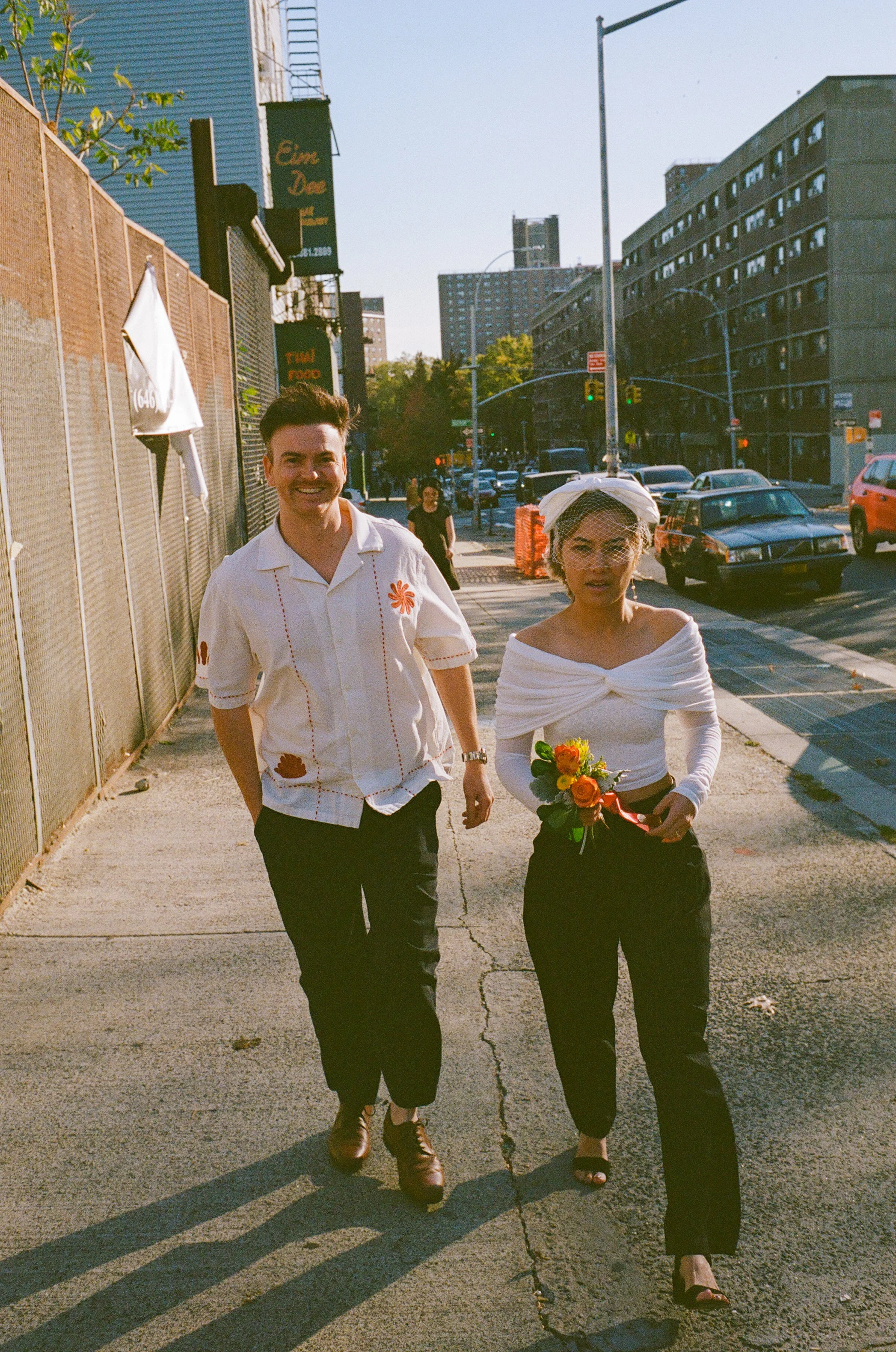 A man and woman walking on the sidewalk in an urban area, with the woman holding a small bouquet of orange roses, the man smiling, and cars parked and driving in the background.