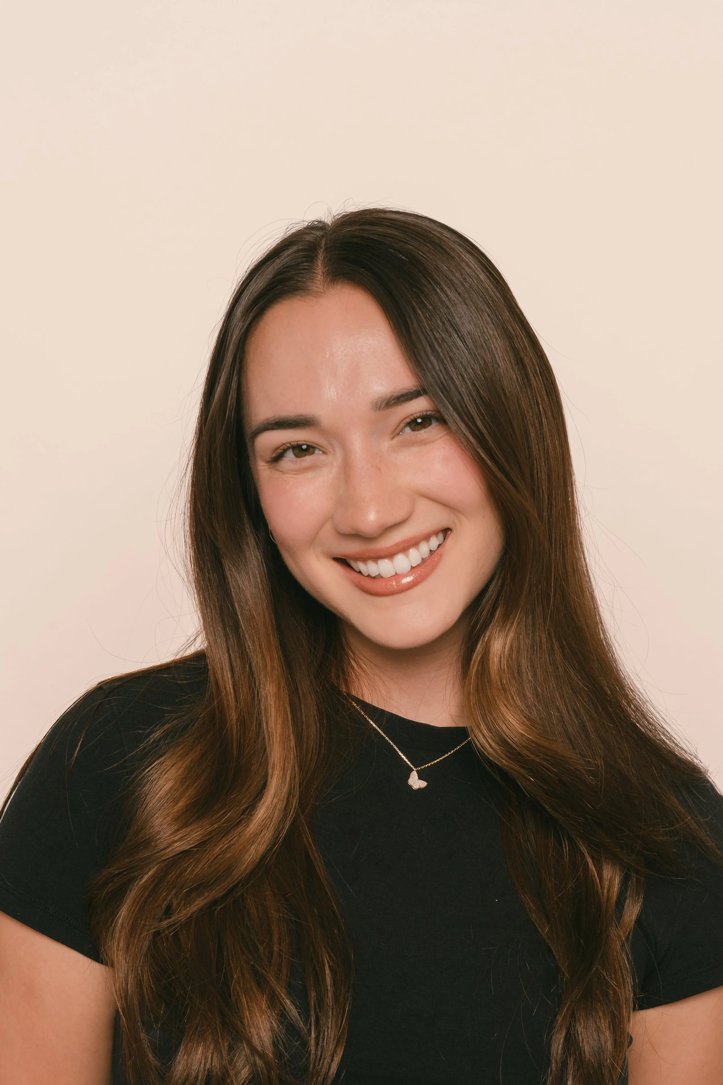 A young woman with long brown hair smiling, wearing a black top and a necklace with a small pendant, against a plain light background.