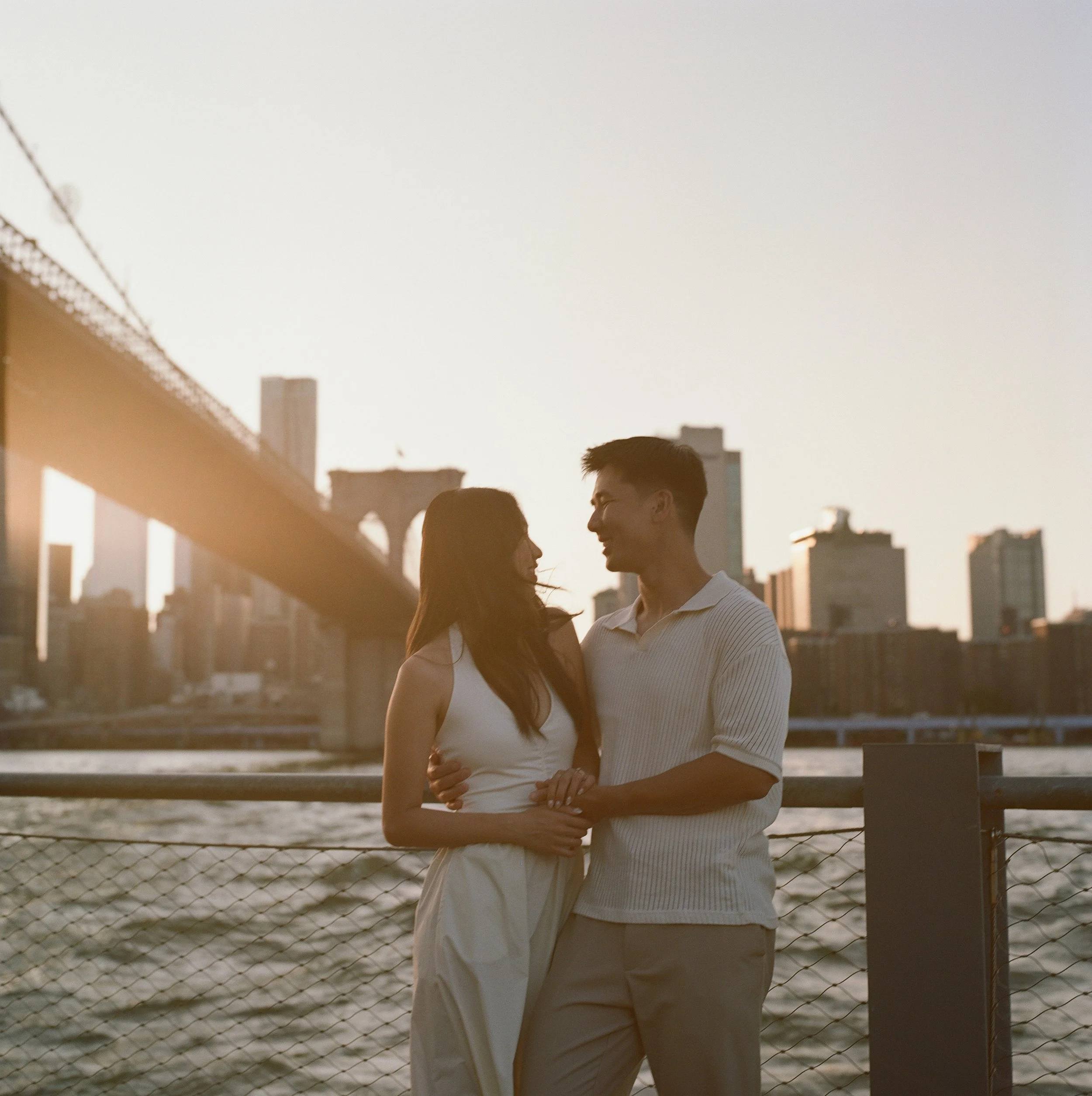 A young couple standing close together by the water with the Brooklyn Bridge and Manhattan skyline in the background during sunset.
