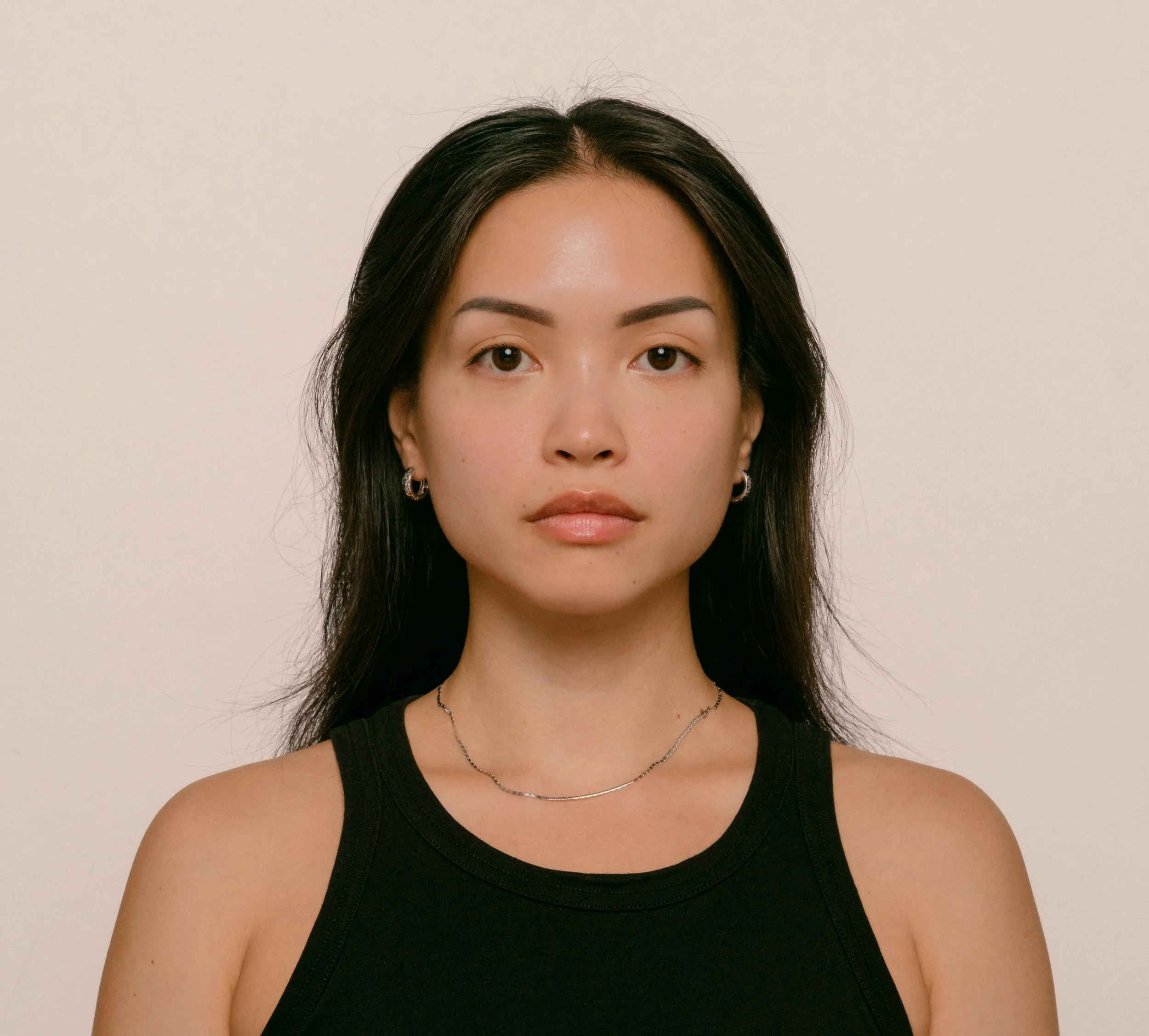 Portrait of a young woman with dark hair, wearing a black tank top, jewelry, and looking directly at the camera against a plain background.