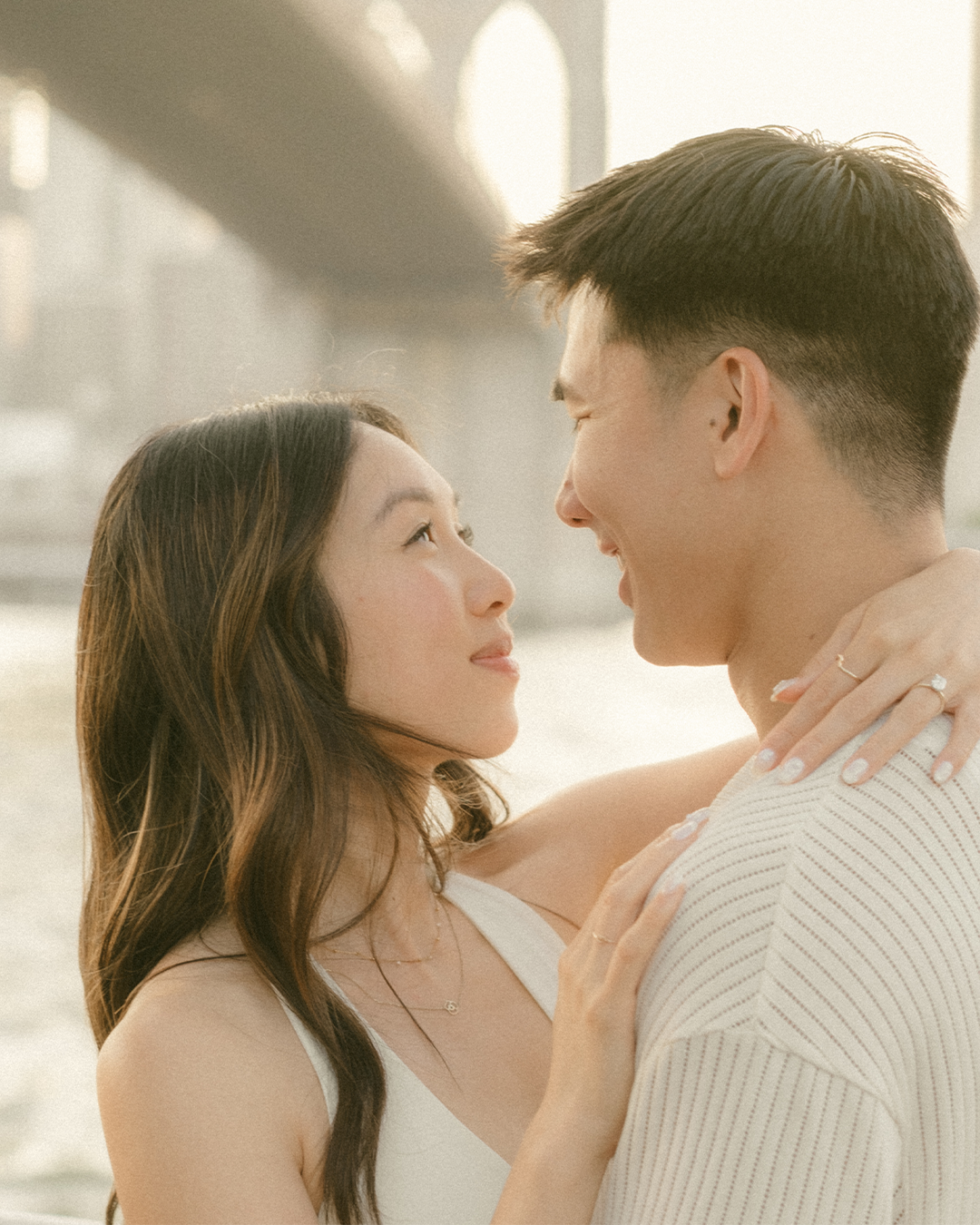A young couple looking at each other lovingly, with a city bridge in the background.