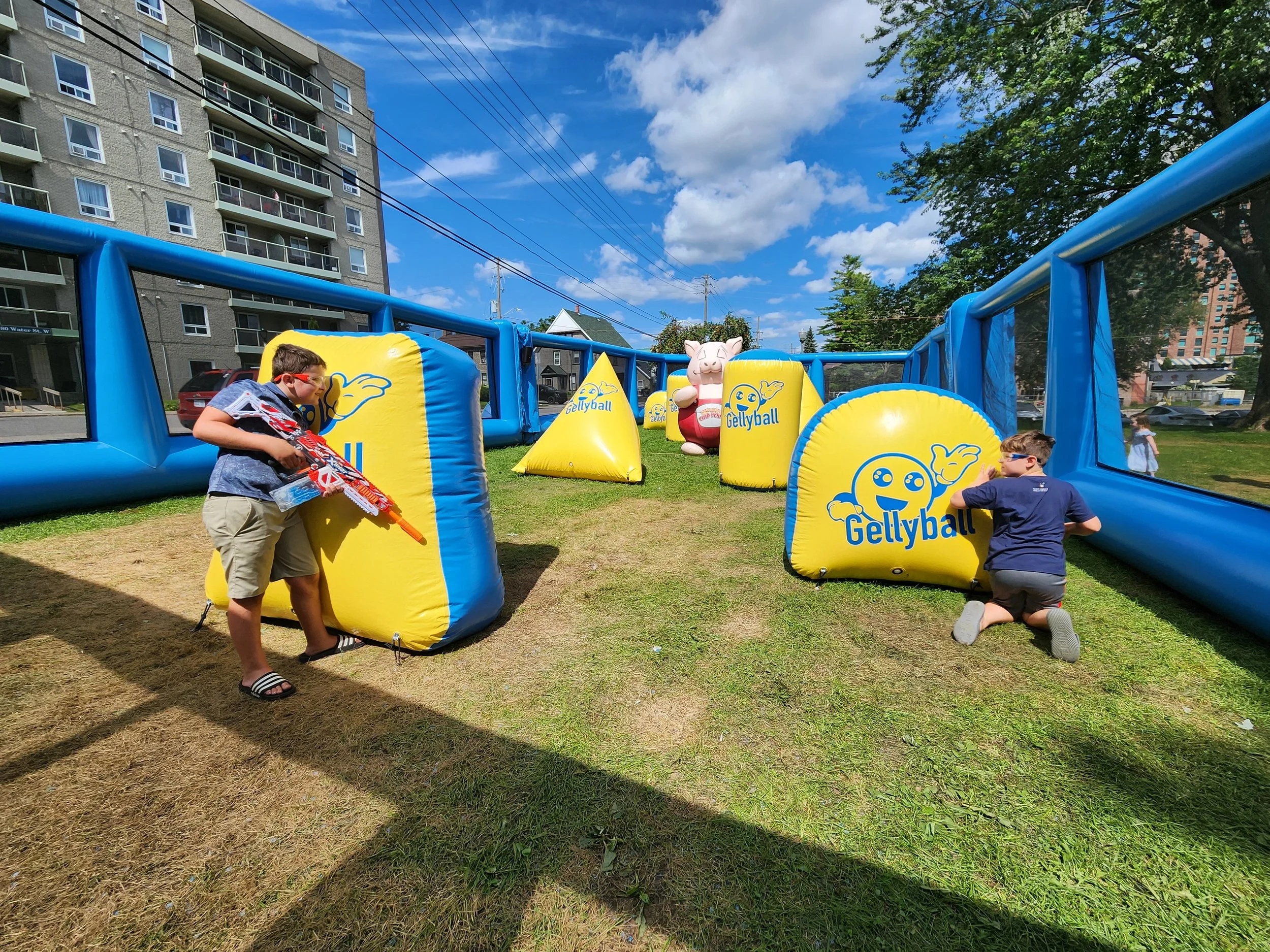 Children playing in a yard with inflatable Gellyball targets and pig character, under a bright blue sky with scattered clouds, nearby buildings and trees.