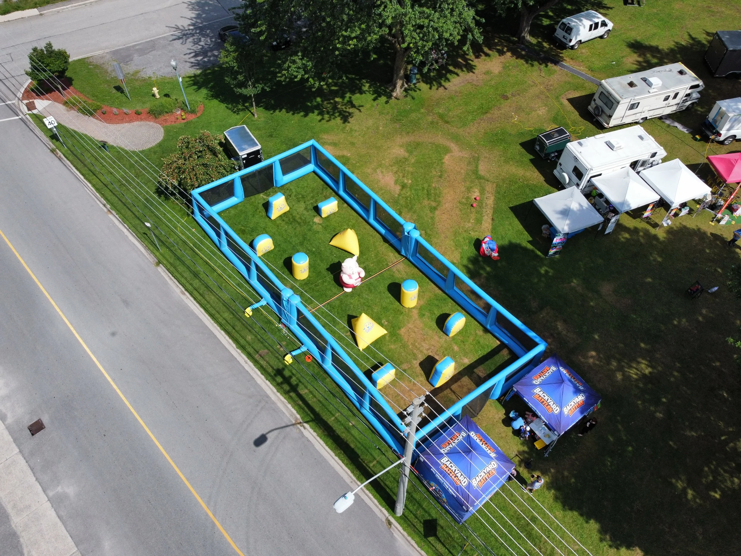 An aerial view of a grassy park area with tents, inflatable attractions, and trailers. There are trees, a sidewalk, and a street with a curb and a fire hydrant visible.