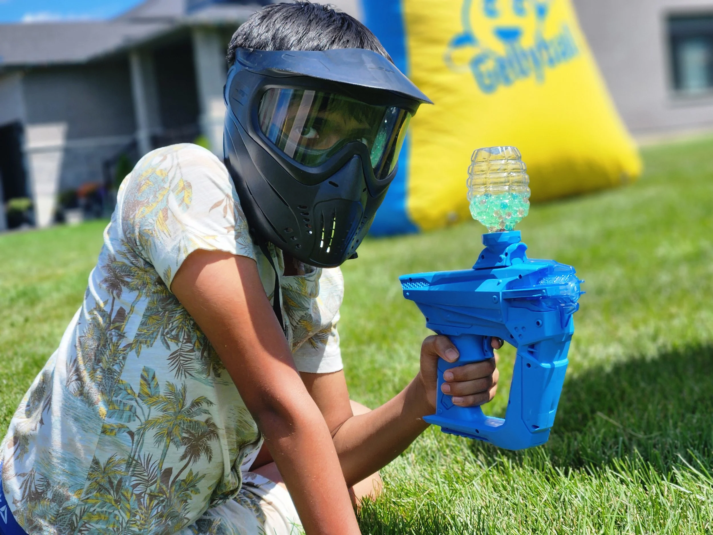 A child in a floral shirt with gray hair playing outdoors with a blue toy gun and wearing a dark face shield, sitting on green grass, with a large yellow and blue inflatable structure in the background.