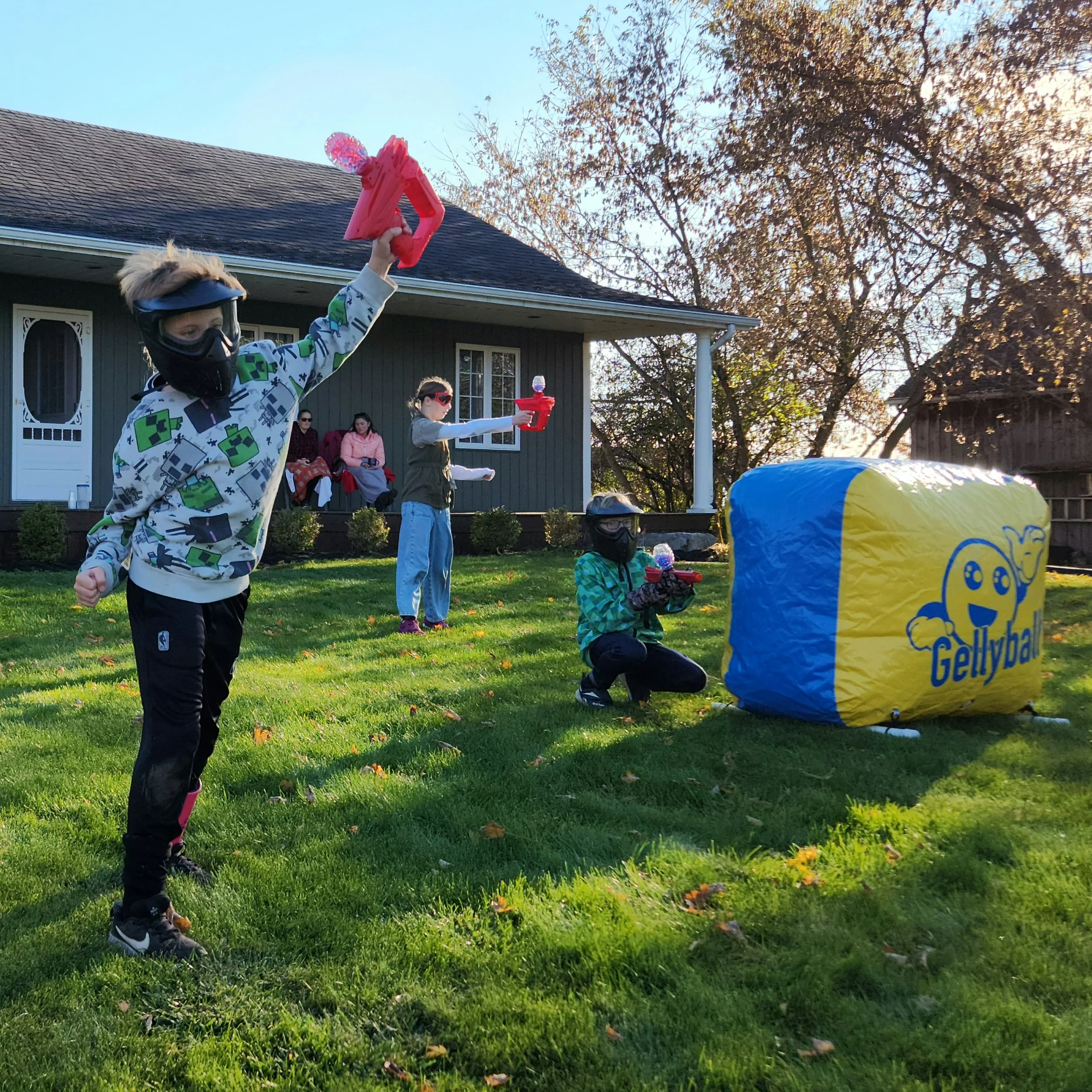 Children playing with water guns outdoors in a backyard with a house, trees, and a grassy lawn, during late afternoon.