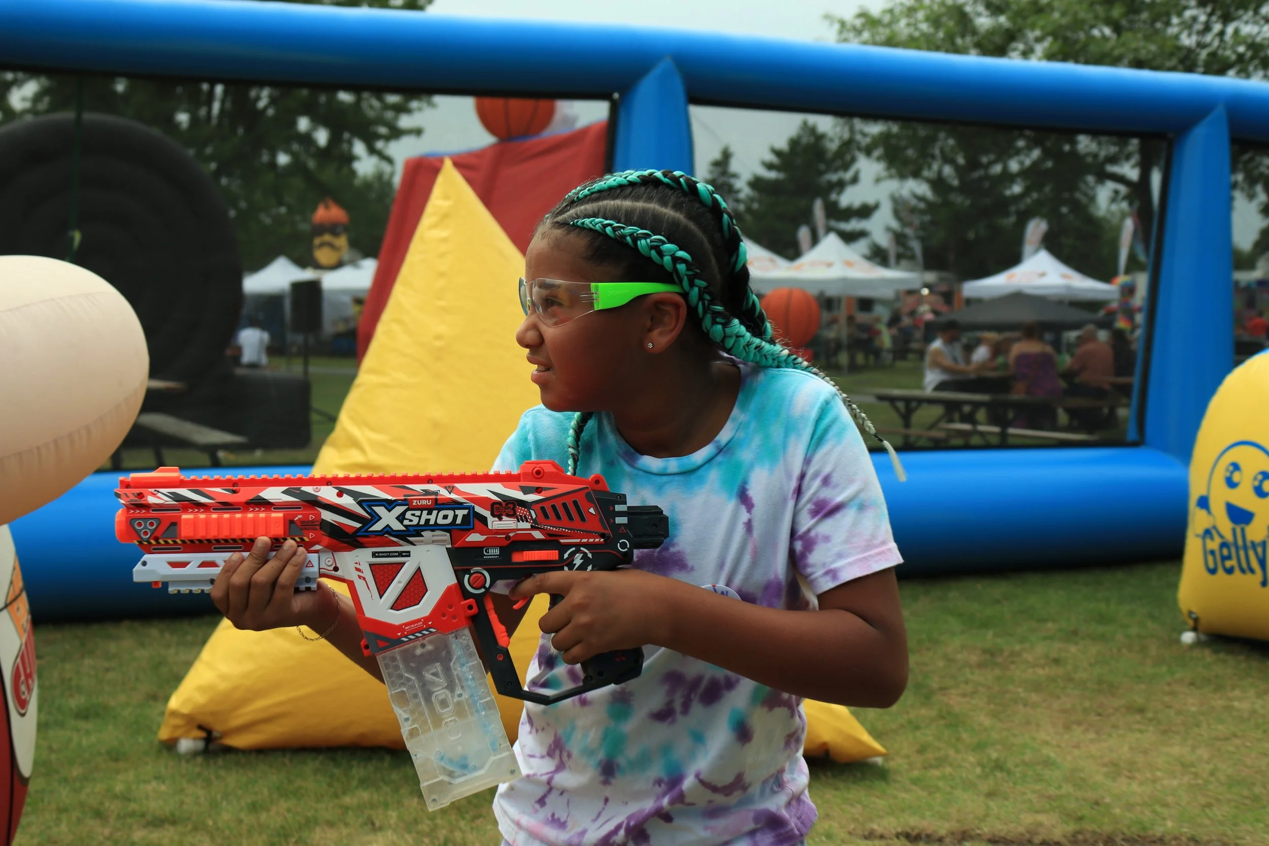 A young girl with braided hair, wearing safety glasses and a tie-dye shirt, holding a large nerf-style toy gun at an outdoor event, with tents, trees, and people in the background.