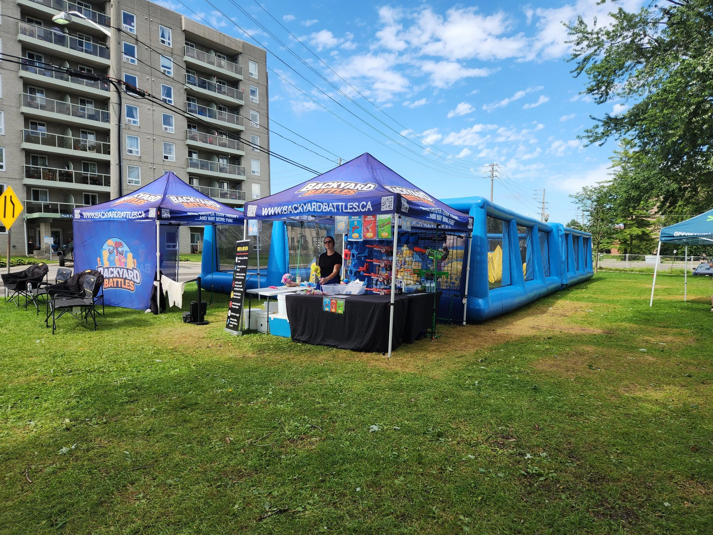 An outdoor event setup with blue tents and a large inflatable structure, with a high-rise residential building in the background and a clear blue sky.