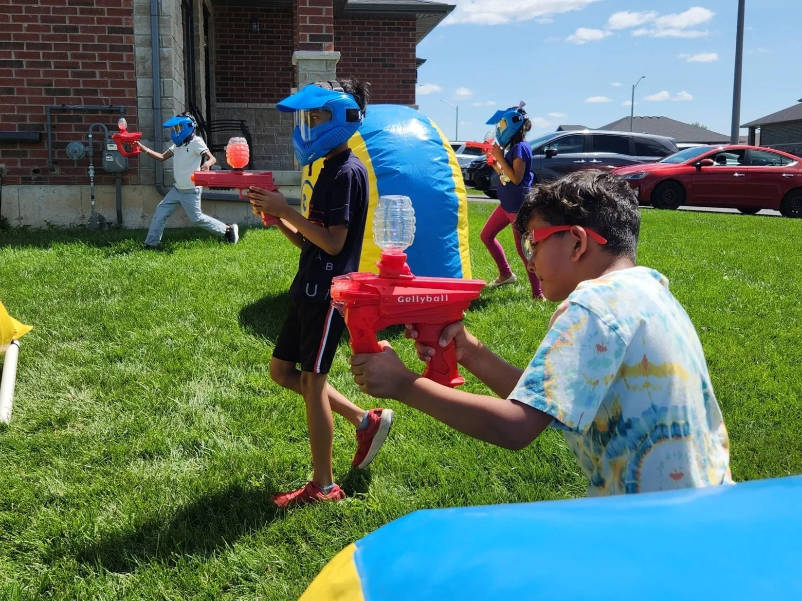 Children playing outdoors with water guns, wearing helmets and protective glasses on a grassy lawn, with a house, parked cars, and blue sky in the background.