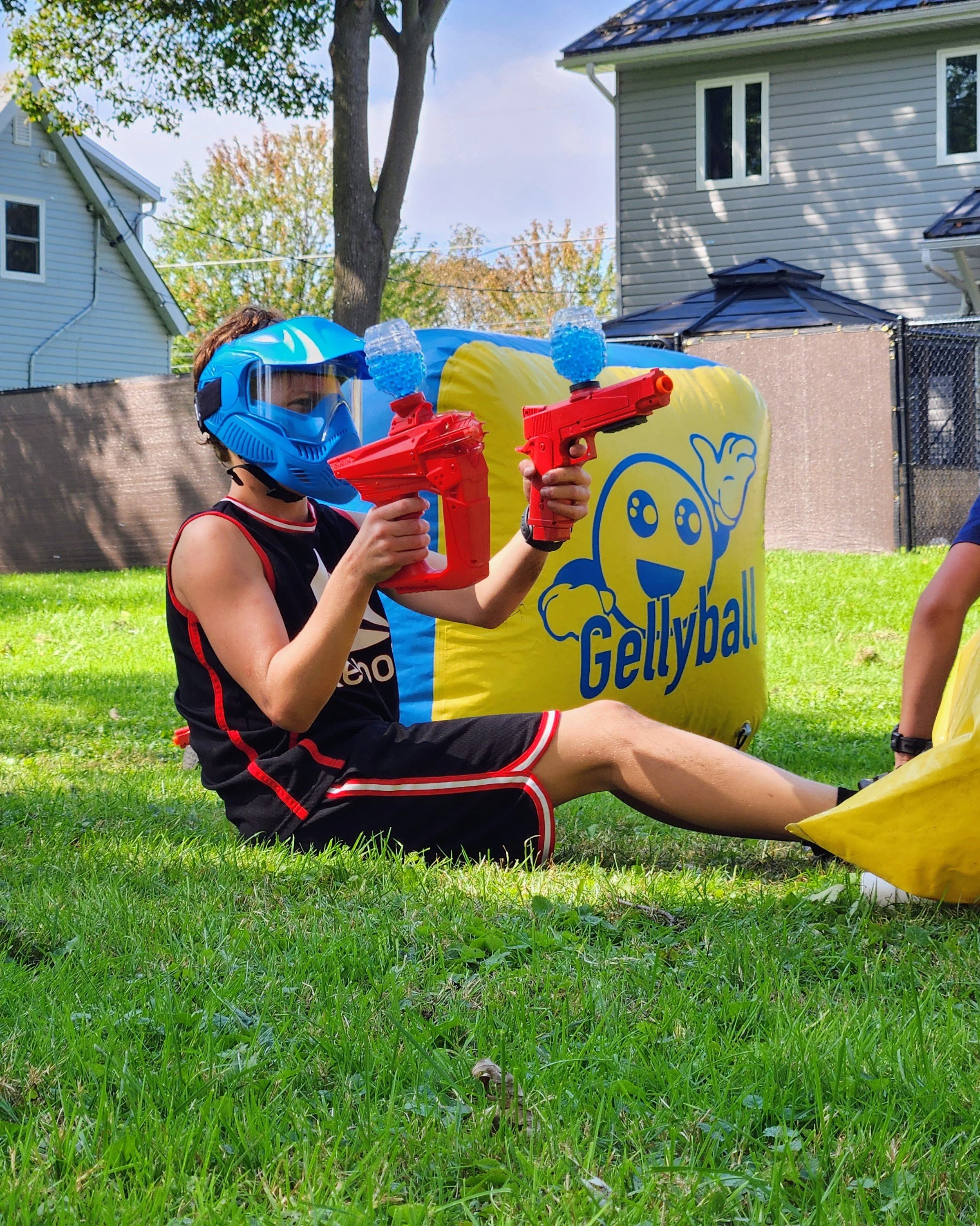 A young person sitting on grass outdoors, wearing a blue helmet and holding two red GellyBall blasters, with a yellow inflatable bunkerl nearby. They are in a yard with houses and trees in the background.