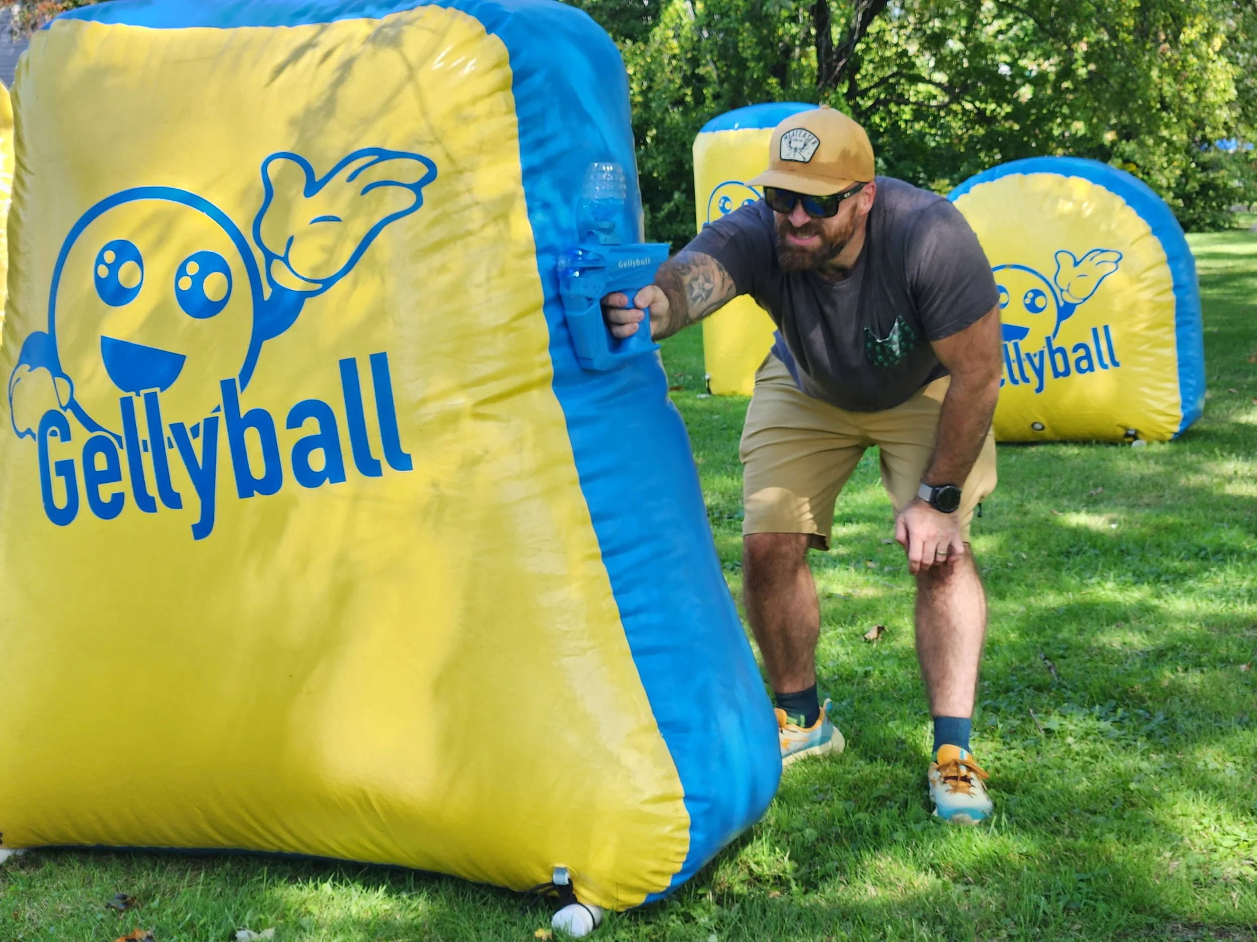 A man wearing sunglasses, a cap, and athletic clothing is crouching behind a yellow and blue inflatable obstacle labeled 'Gellyball' outdoors on a grassy area with trees in the background. The obstacle has a cartoon face and waving hand design.