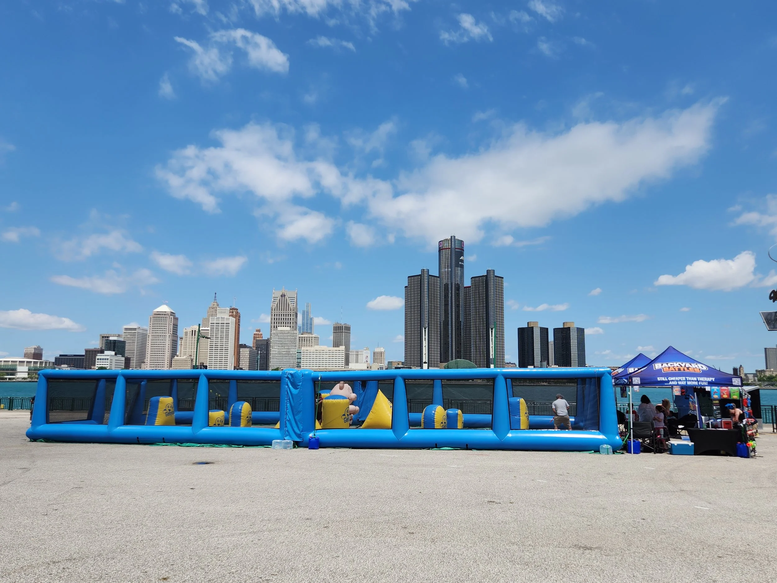 A city skyline with tall buildings under a blue sky with scattered clouds. In the foreground, there is a blue inflatable bounce house and tents, with people gathered nearby.