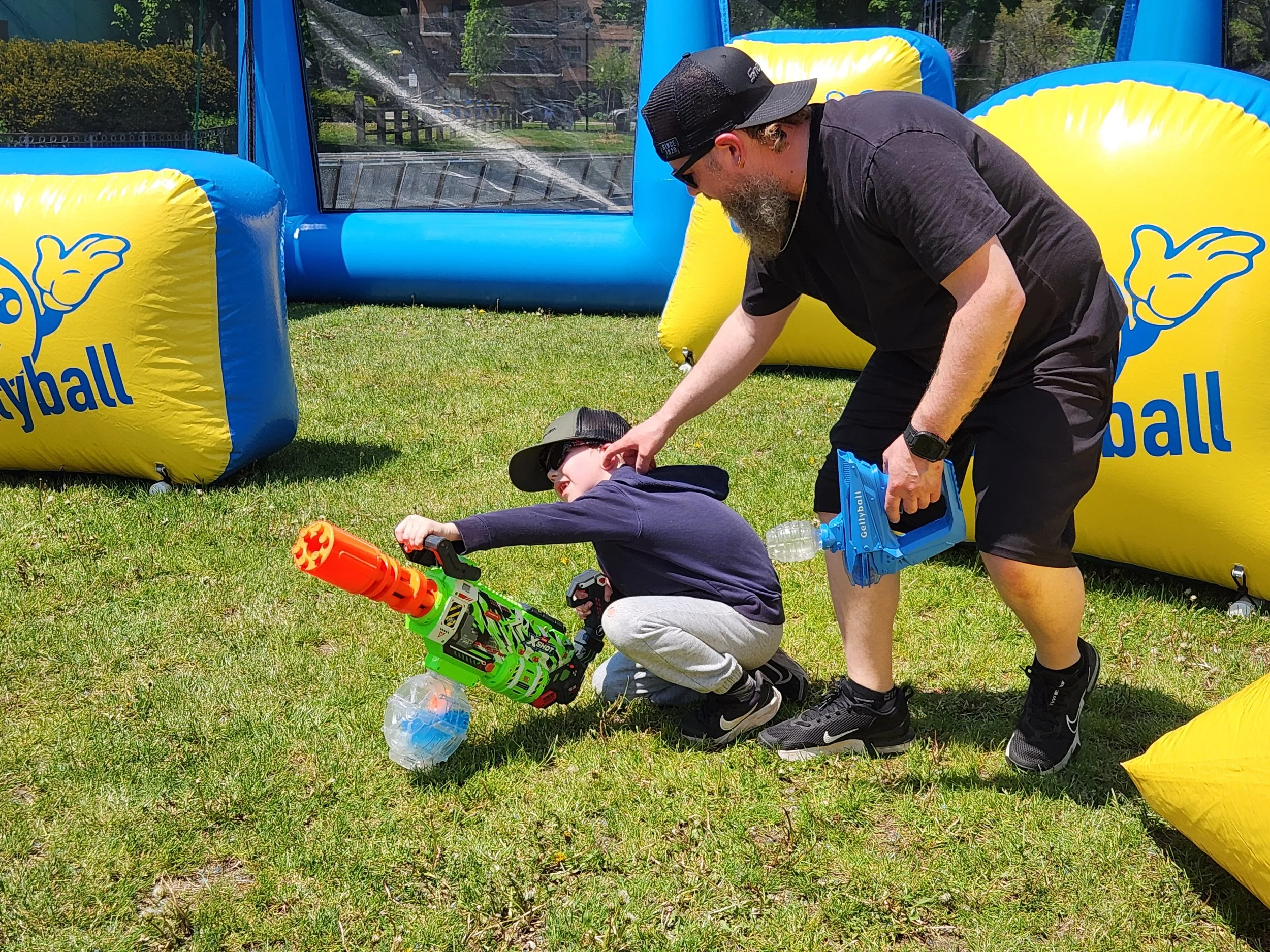 A man with a beard, wearing a black shirt, shorts, cap, and sunglasses, helps a young boy in a black jacket and gray pants with a brightly colored toy gun at an outdoor inflatable bounce house decorated for a summer event.
