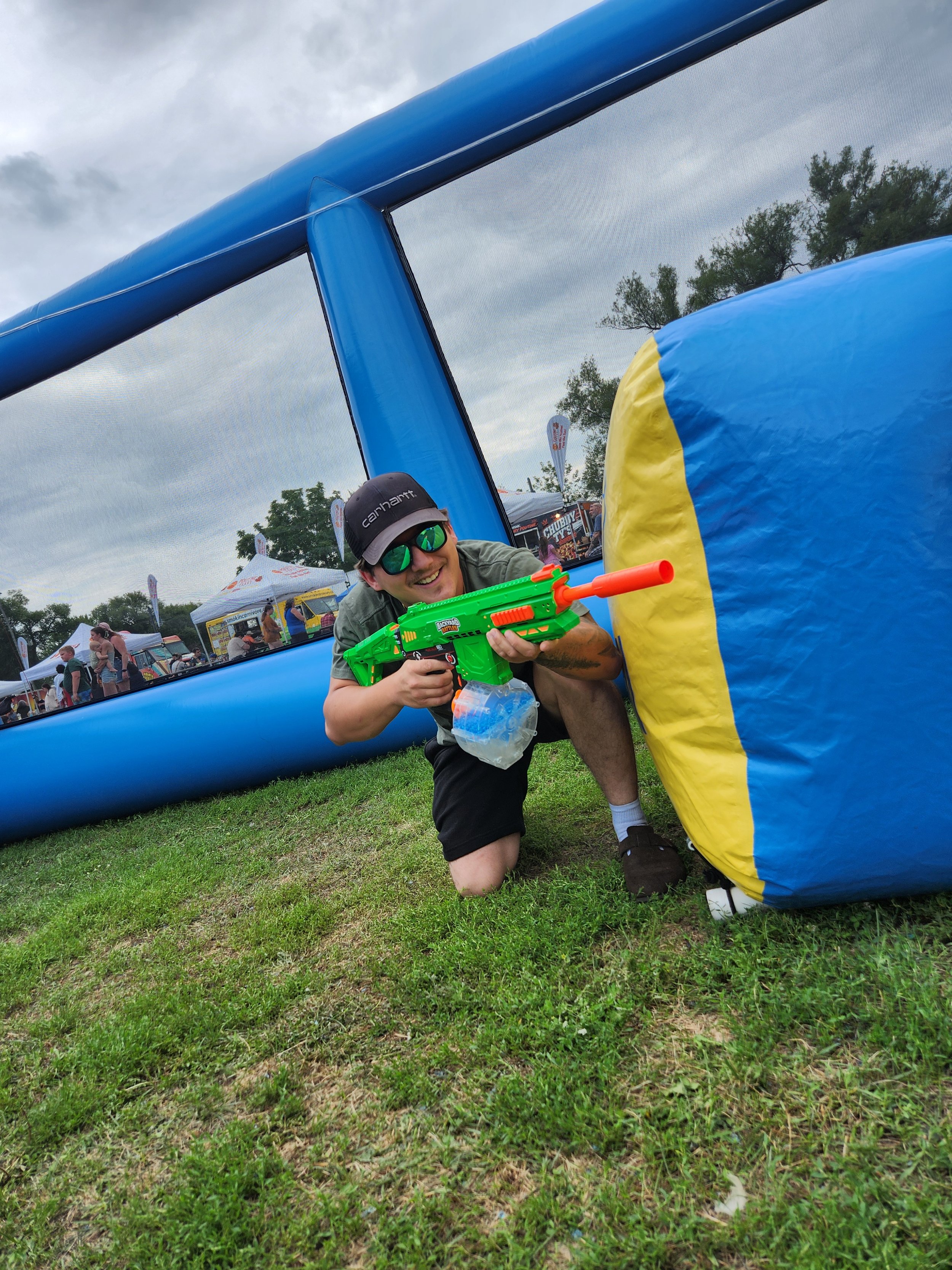 Man in sunglasses holding a water gun and crawling on the grass at an outdoor event with a large inflatable structure.