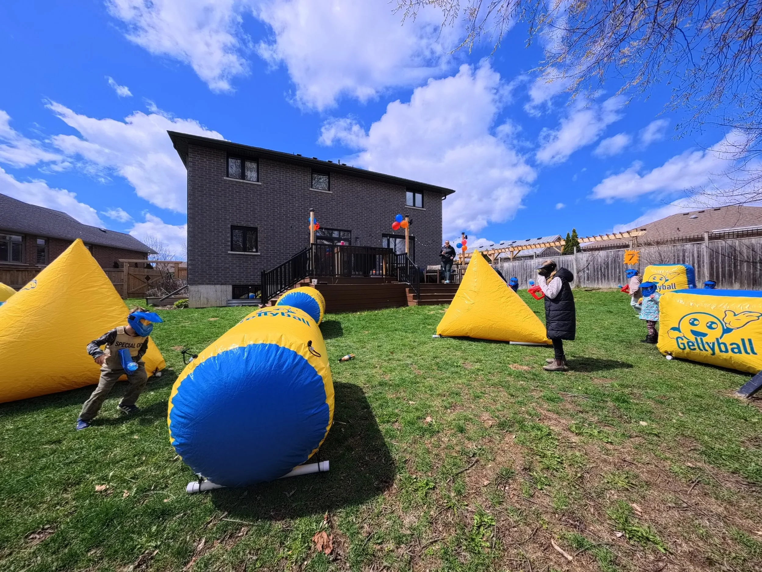 Children and adults playing Gellyball with inflatable barriers in a backyard on a sunny day.