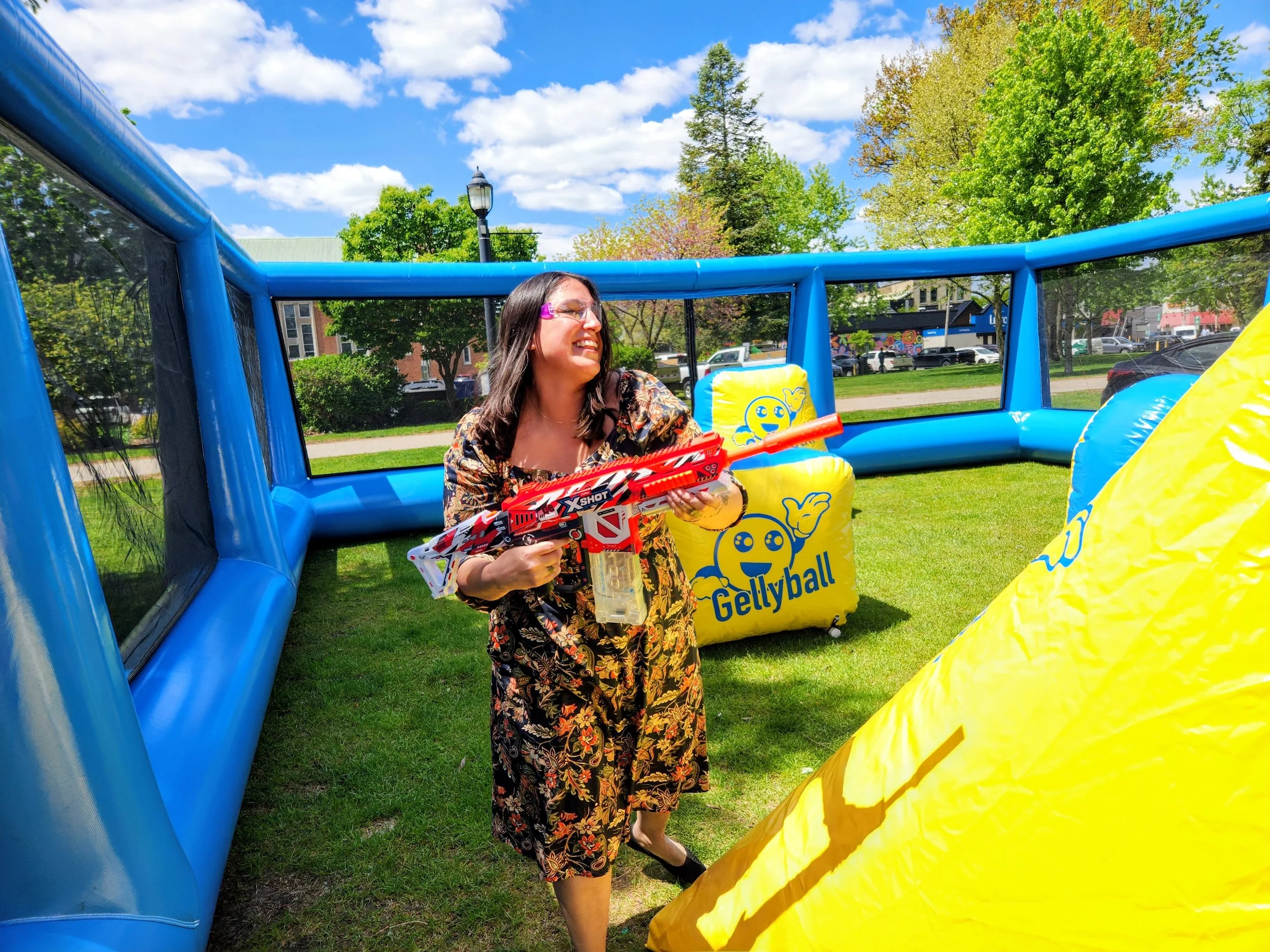 Woman holding a toy gun inside a blue bounce house at an outdoor event with trees and a grassy area.