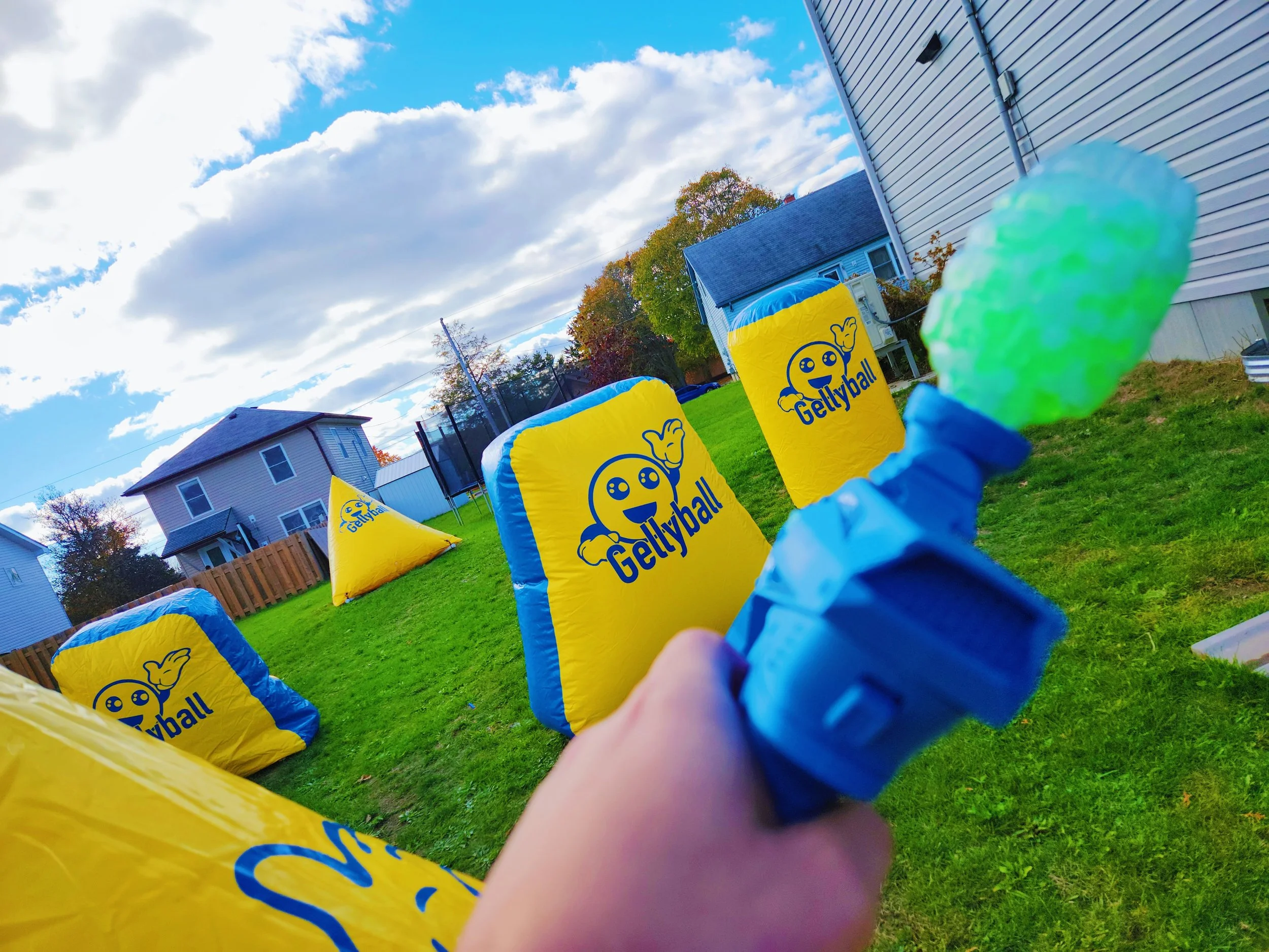Backyard with a blue house, trees with fall foliage, and inflatable gym mats with Gettyball logo, under a partly cloudy sky.