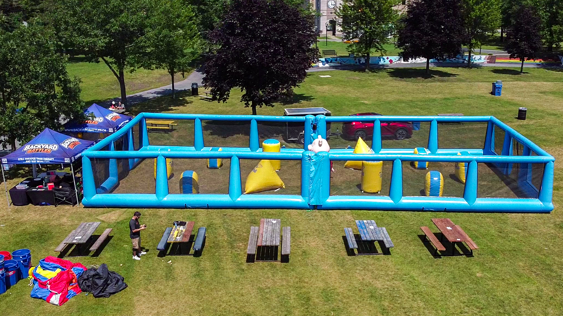 An outdoor park with a large blue inflatable obstacle course, a white unicorn-shaped bounce house, and a tent labeled Backyard Battles. There are picnic tables, a person standing with a phone, and several blue, red, and black bean bags on the grass.