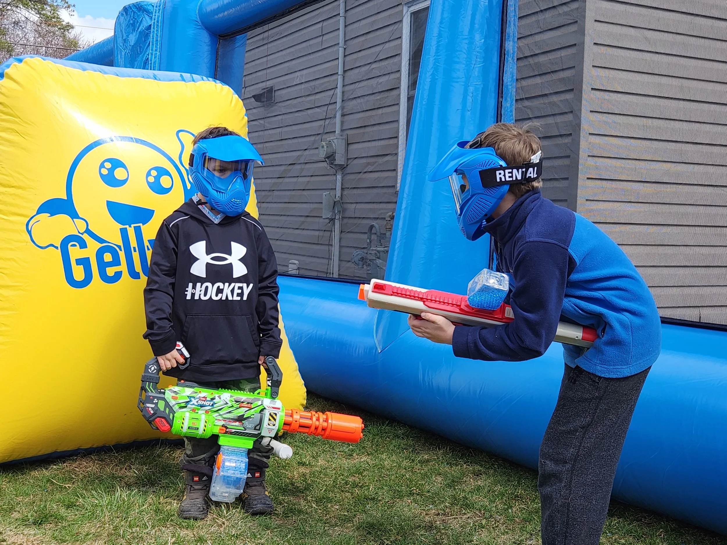 Two children wearing blue helmets and face masks playing outdoors near an inflatable blue and yellow structure. One child is holding a toy blaster, and the other appears to be talking to an adult holding a toy. There is a yellow inflatable structure 