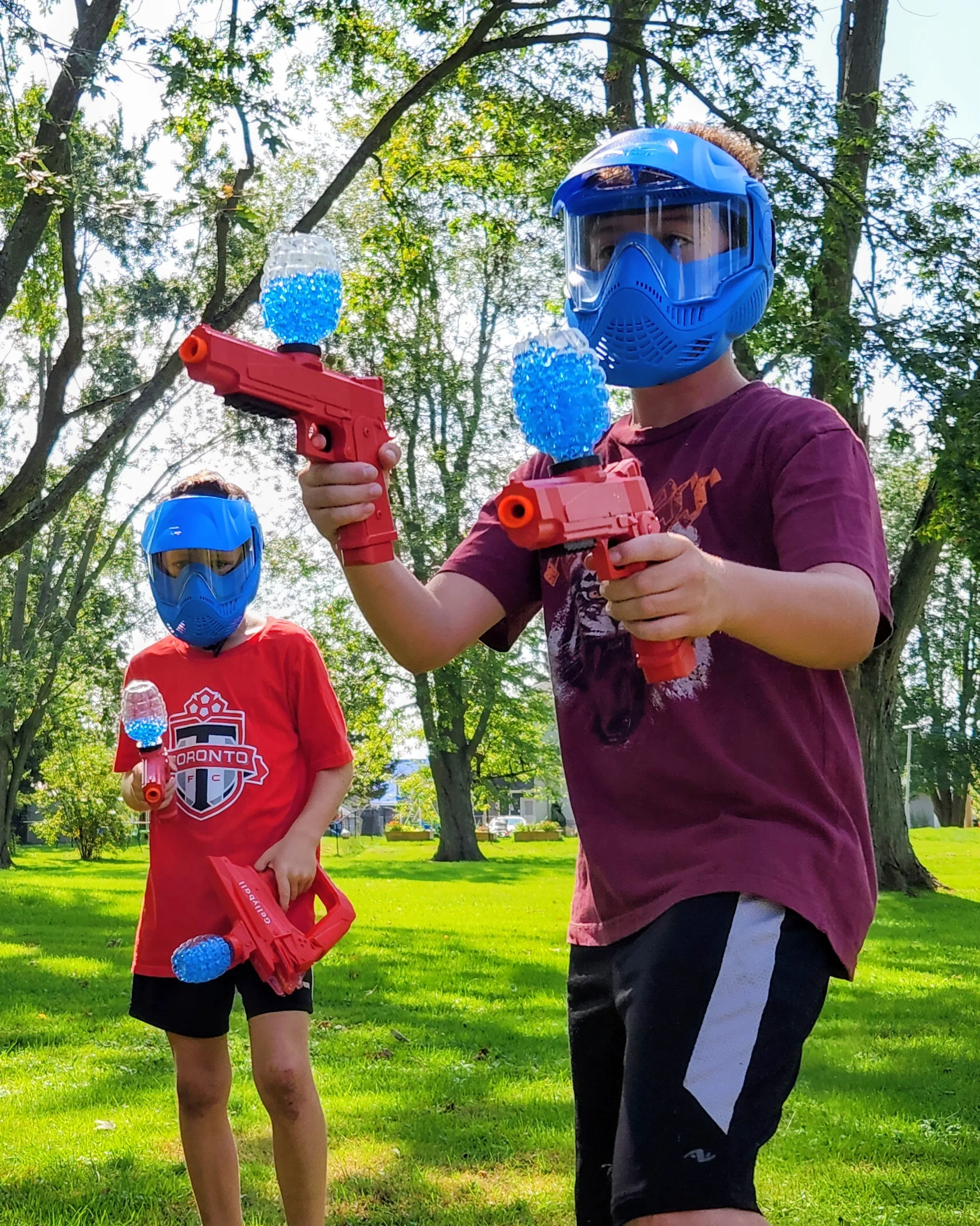 Two children wearing blue shield helmets holding red and blue toy guns and water balloons outdoors in a park with trees and grass.