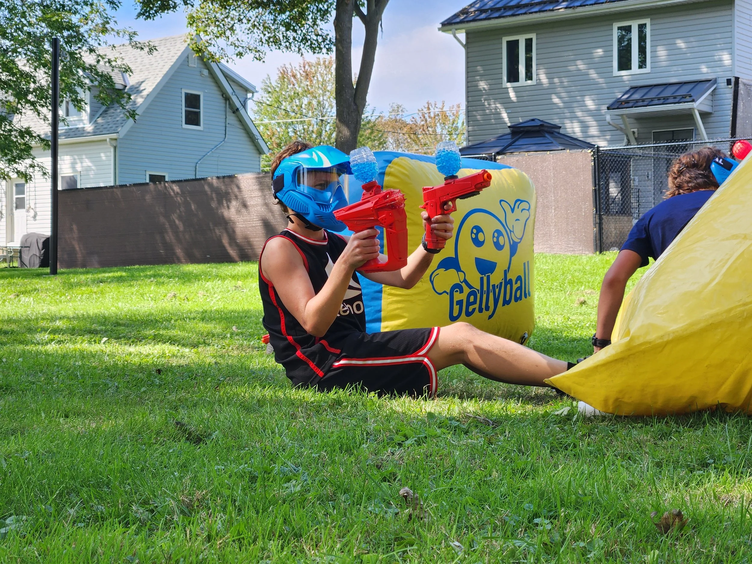 Child wearing a blue helmet and black shirt, sitting on grass, holding two toy water guns, with a yellow colt-style bounce house in a backyard.