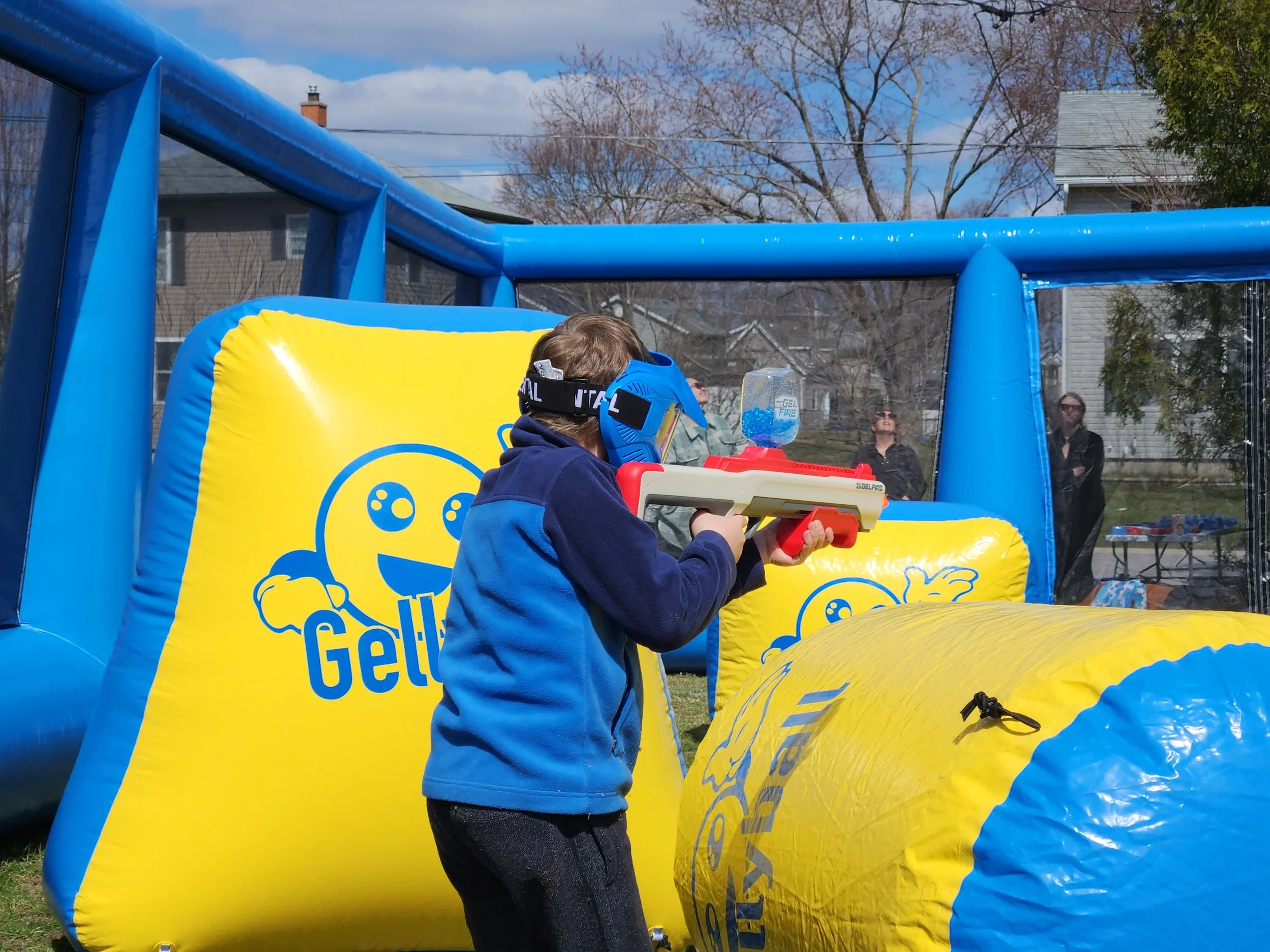 Child wearing a blue jacket, helmet, and goggles aiming a water gun at an outdoor inflatable Gel Blaster arena with blue and yellow branding. Several adults are seen watching in the background on a clear day.