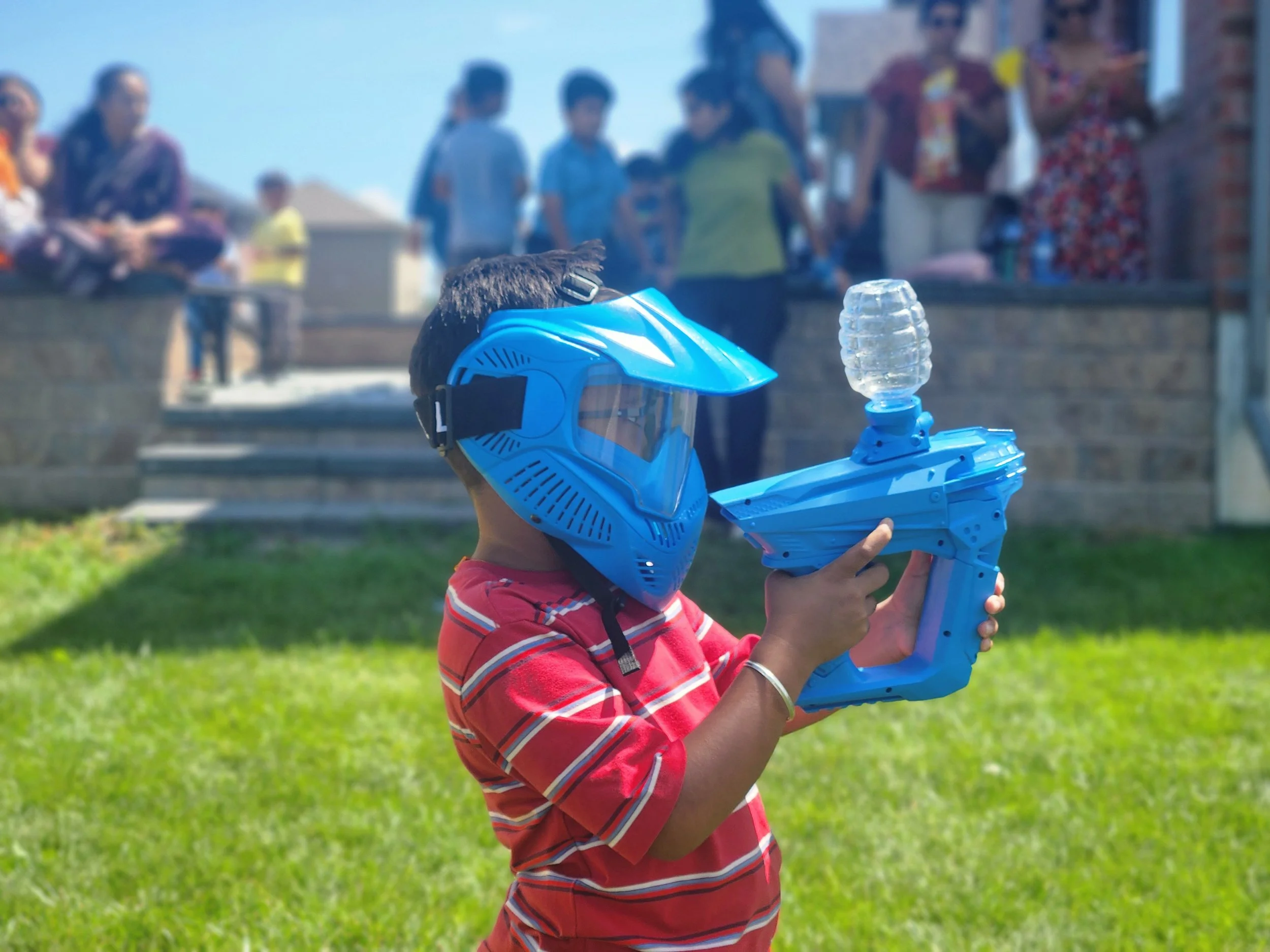 Child wearing a blue helmet and goggles, holding a toy water gun outdoors on a sunny day, with people standing on a ledge in the background.