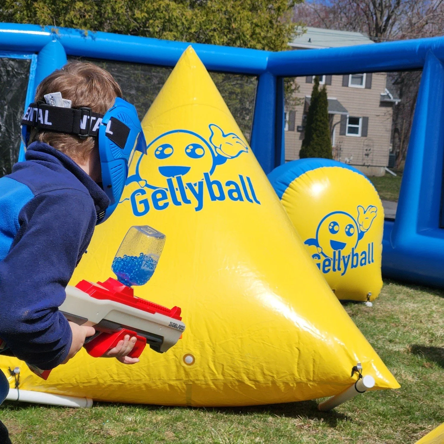 Child wearing a blue helmet and blue jacket holding a toy gun, aiming at yellow inflatable obstacles with 'Gellyball' logo at an outdoor event.