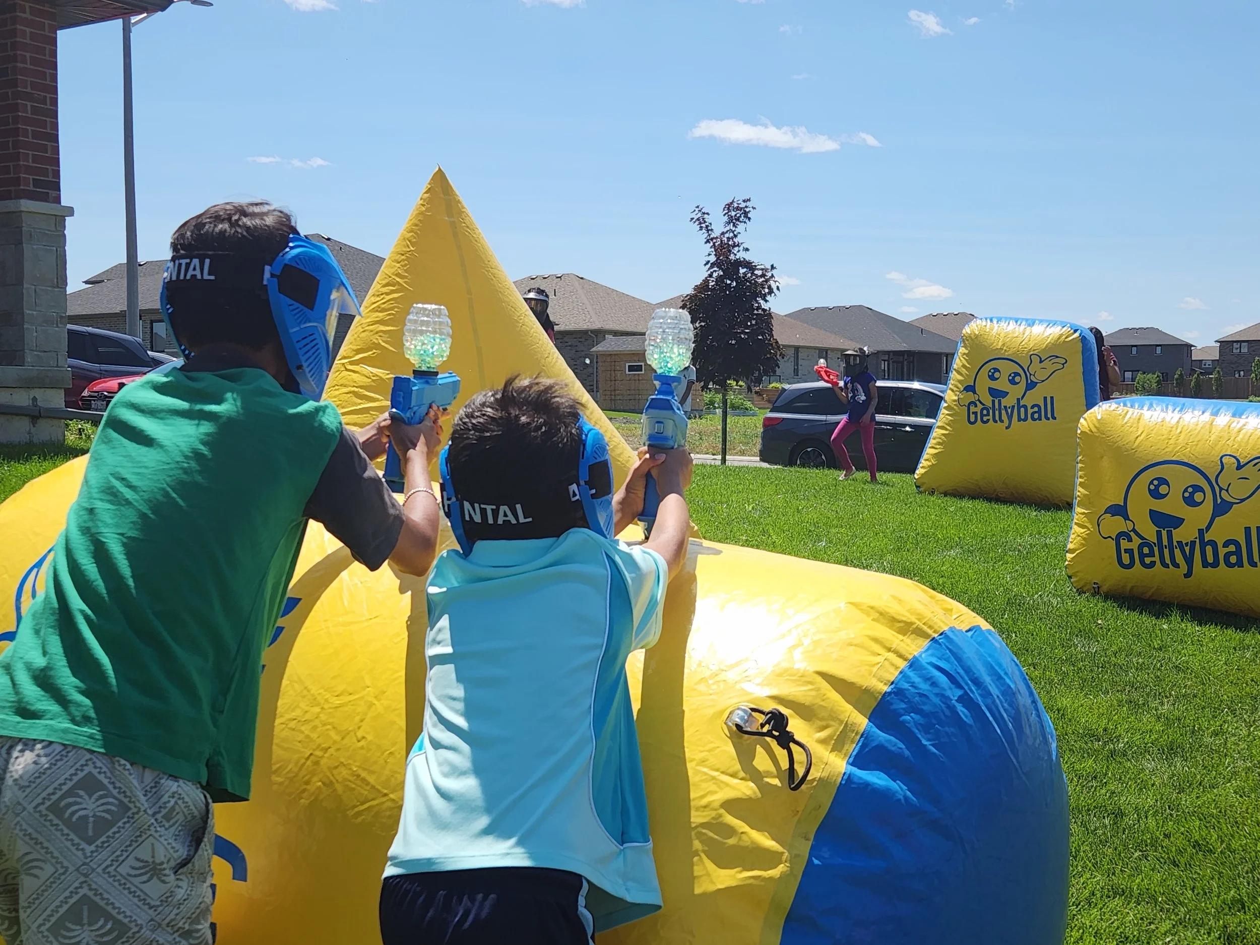 People participating in a bubble soccer game outdoors on a grassy lawn, with inflatable barriers and goggles, under a clear blue sky.