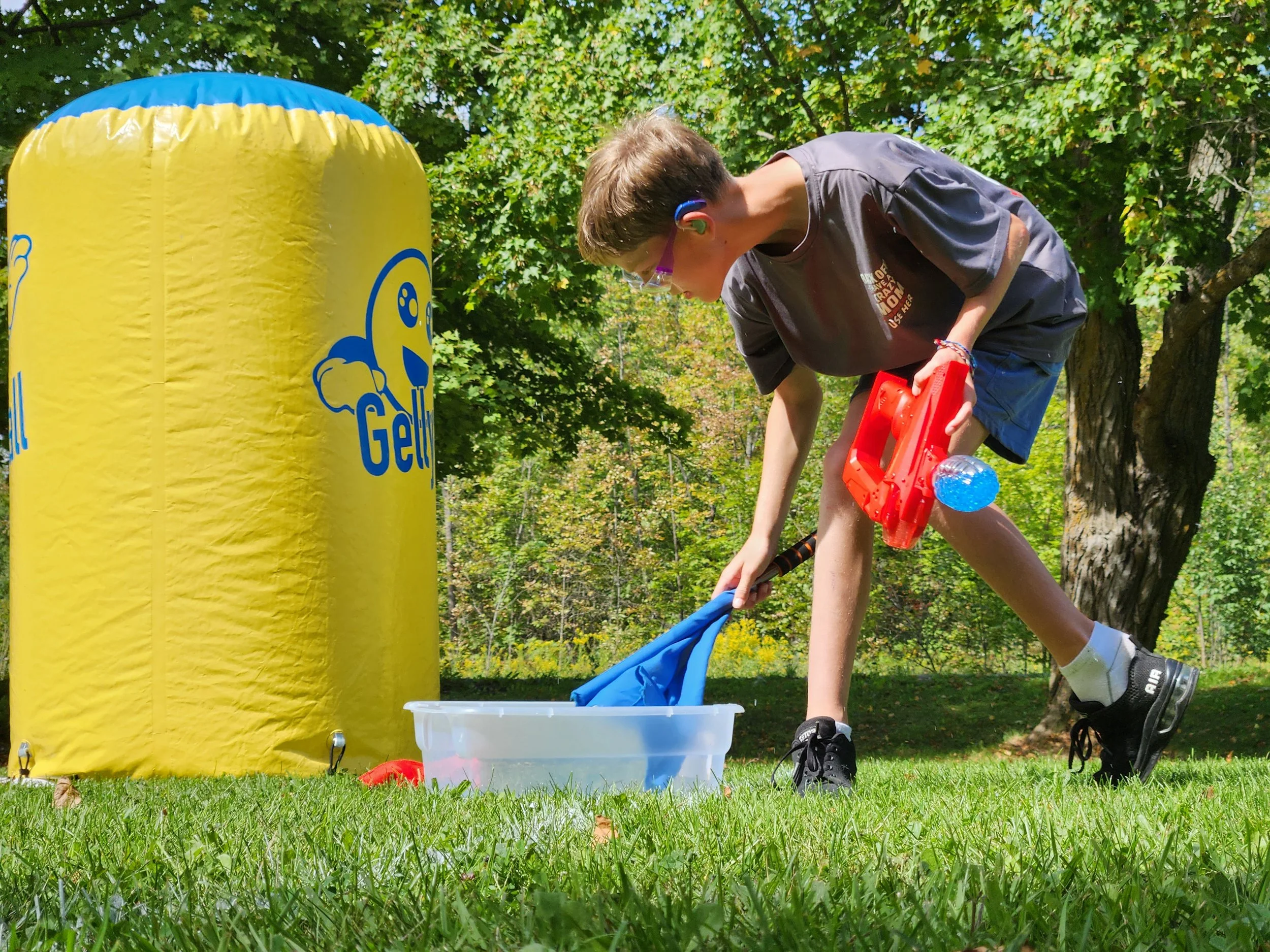 Child with brown hair wearing glasses, black t-shirt, blue shorts, and black sneakers playing outdoors with a water gun near a yellow inflatable structure with a cartoon character and green trees in the background.
