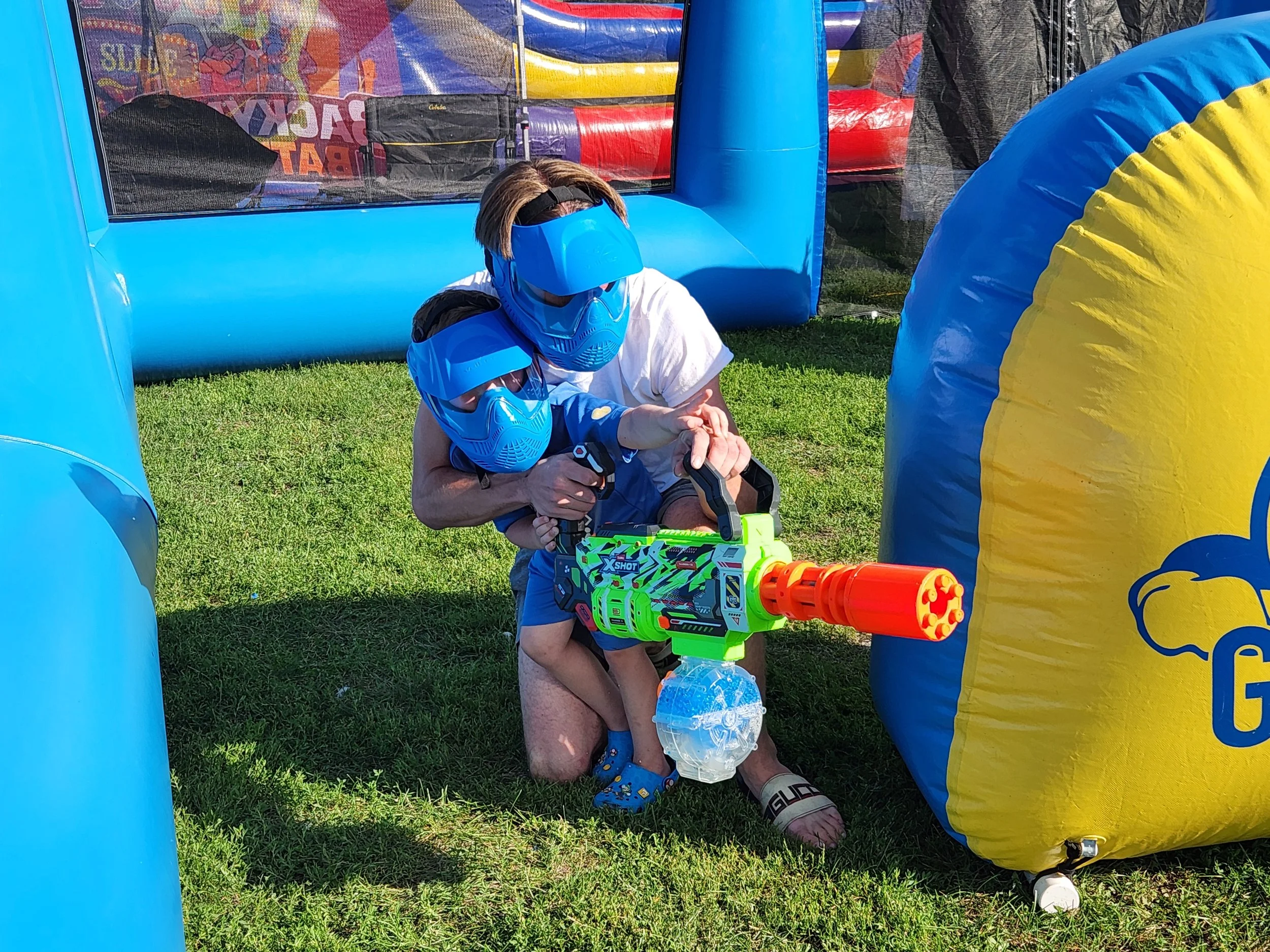 A young child and an adult, both wearing blue protective masks and goggles, are playing with a toy water gun inside an inflatable bounce house at an outdoor event. The bounce house is blue and yellow, and the background shows various colorful inflata