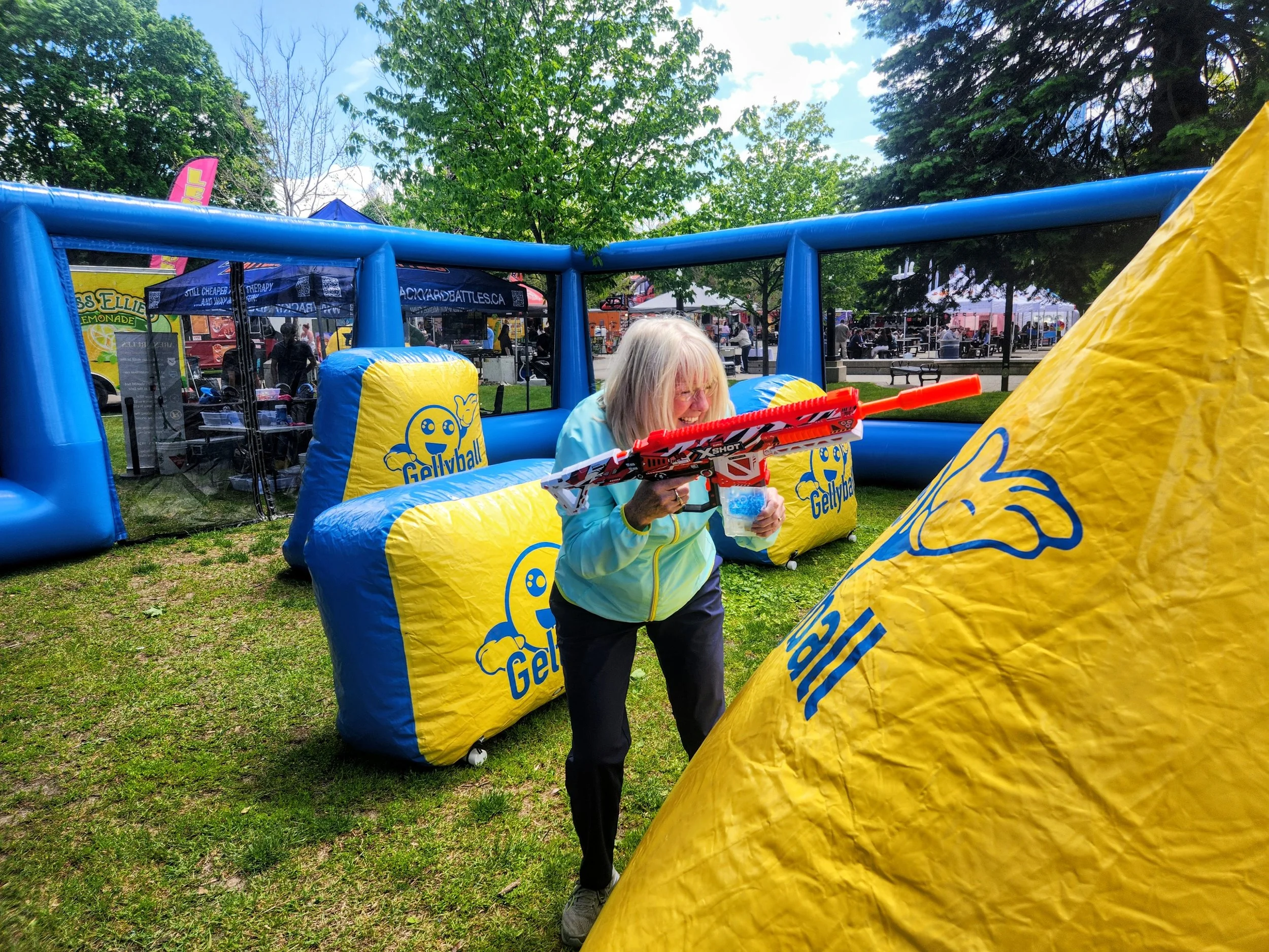An outdoor scene at a fair or festival with a woman holding a toy gun inside an inflatable shooting game. The game is yellow and blue with the logo 'Gellyball.' In the background, there are tents, food stalls, and visitors, with trees and a partly cl