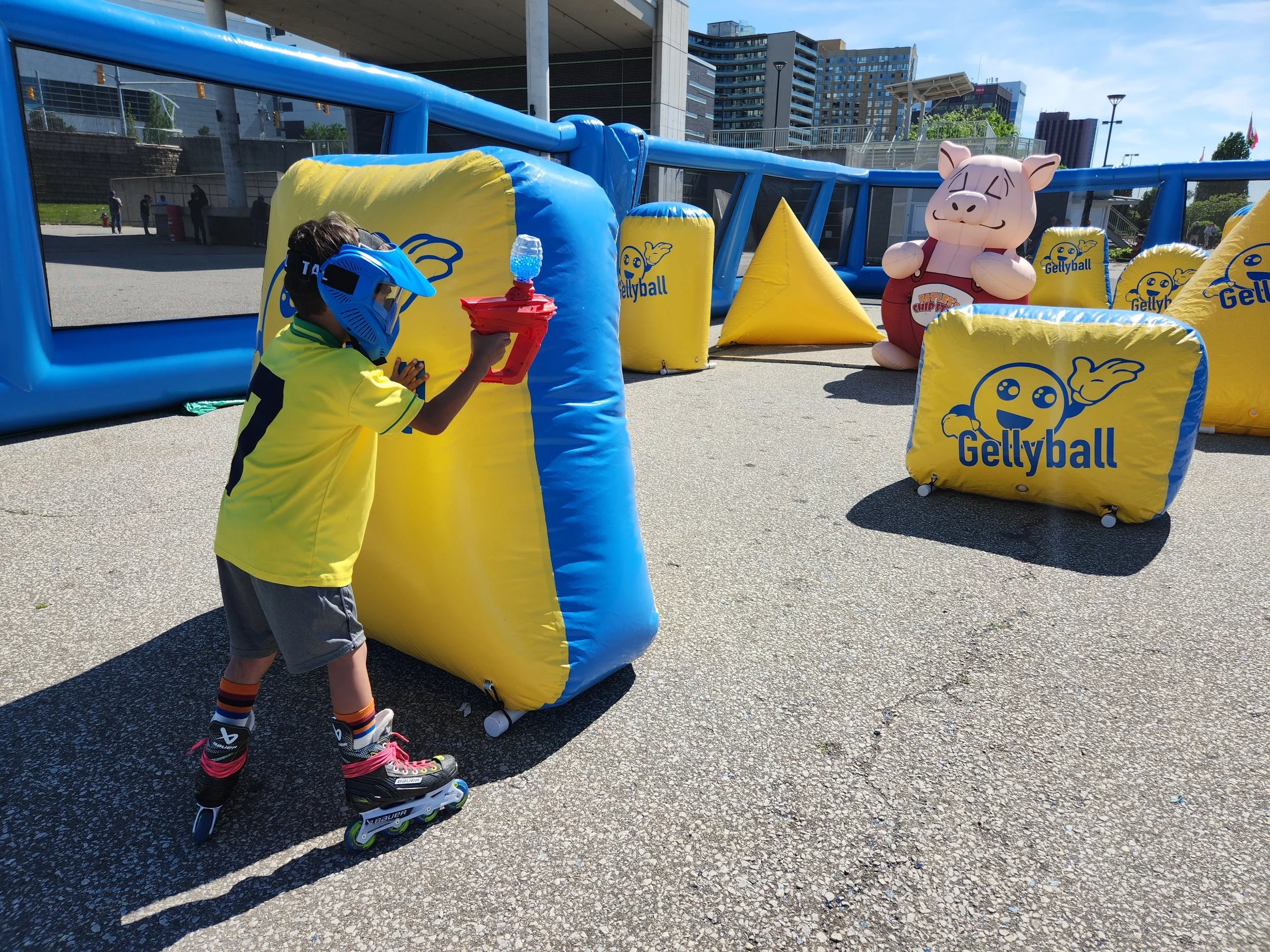 A young boy wearing a blue helmet, yellow T-shirt, and rollerblades is aiming a water gun at a pink inflatable pig mascot during an outdoor event. The scene is set in a city plaza with buildings and a bridge in the background, and inflatable barriers