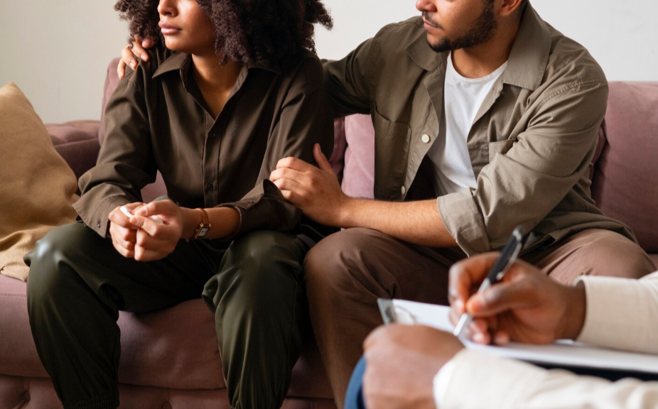 A woman and man sitting close together on a couch during a counseling or therapy session, with a therapist taking notes.
