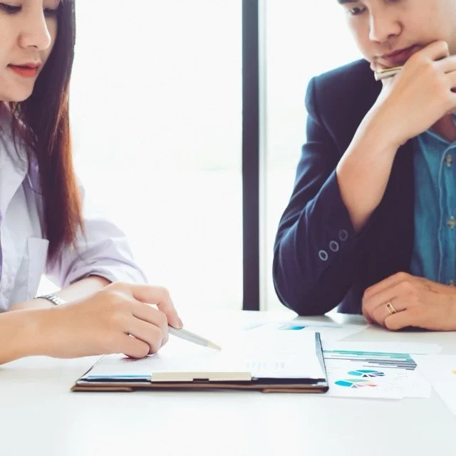Two people sitting at a table, reviewing documents with charts, one woman pointing with a pen and a man holding a pen to his chin, in a well-lit room.