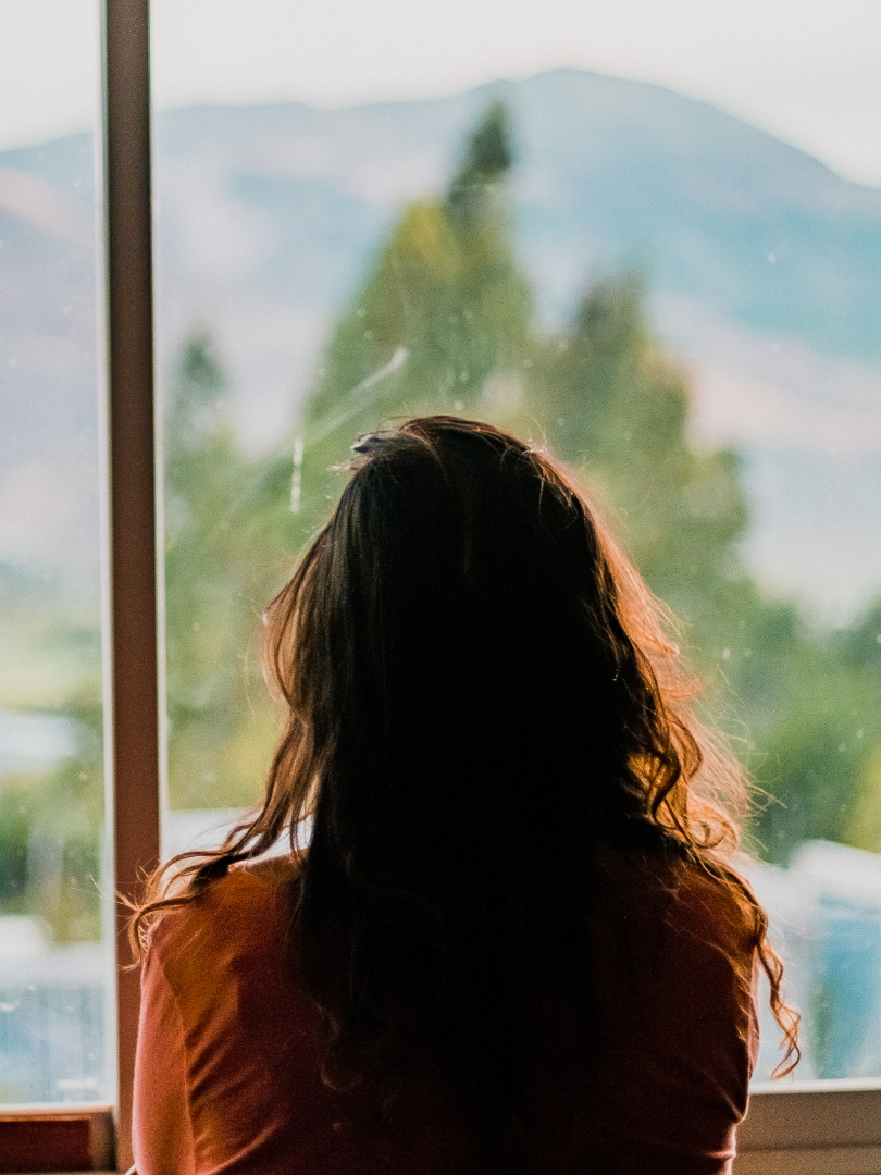 A woman with wavy dark hair looking out a window at a blurry landscape of trees and mountains during daytime.
