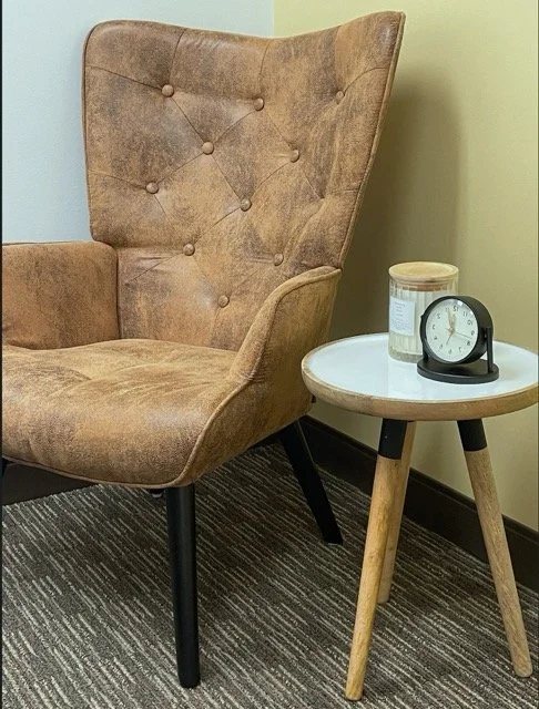 A beige upholstered armchair with tufted buttons next to a small round wooden side table holding a white container and a black analog clock with a round face on a striped rug.
