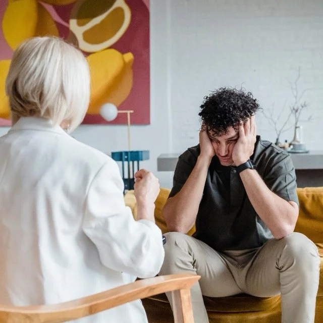 A young man with curly hair and beige pants sitting on a yellow sofa, holding his head in his hands, appearing distressed, while talking with a woman with short blonde hair and a white blazer, who is gesturing with her hand in a cozy living room.