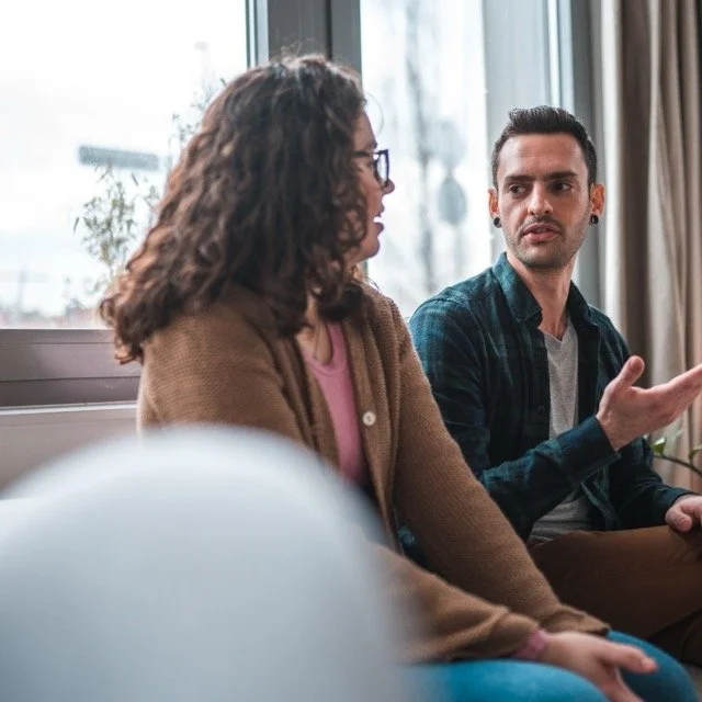 A man and woman sitting and engaging in a conversation in a well-lit room with large windows.