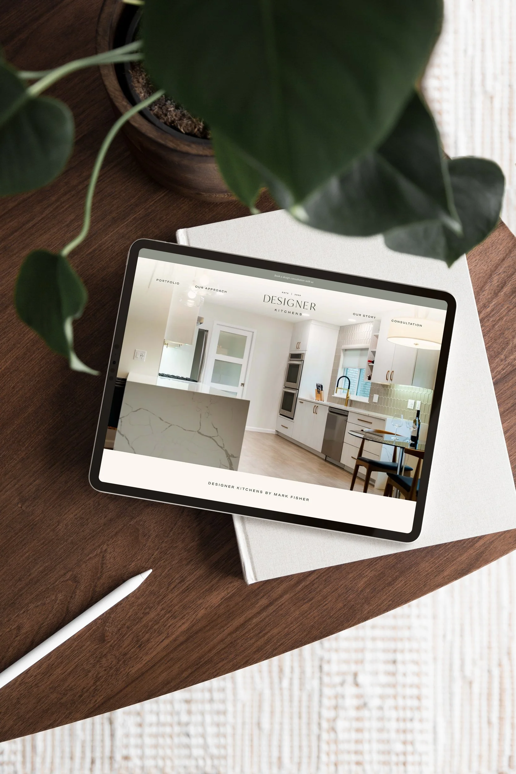 Tablet displaying a website for designer kitchens on a wooden table with a white notebook, pen, and a potted plant nearby.