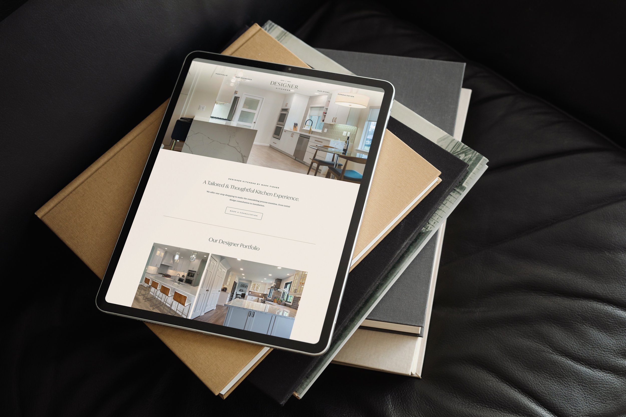 Tablet displaying a website with a modern kitchen interior, resting on a stack of hardcover books on a black leather surface.