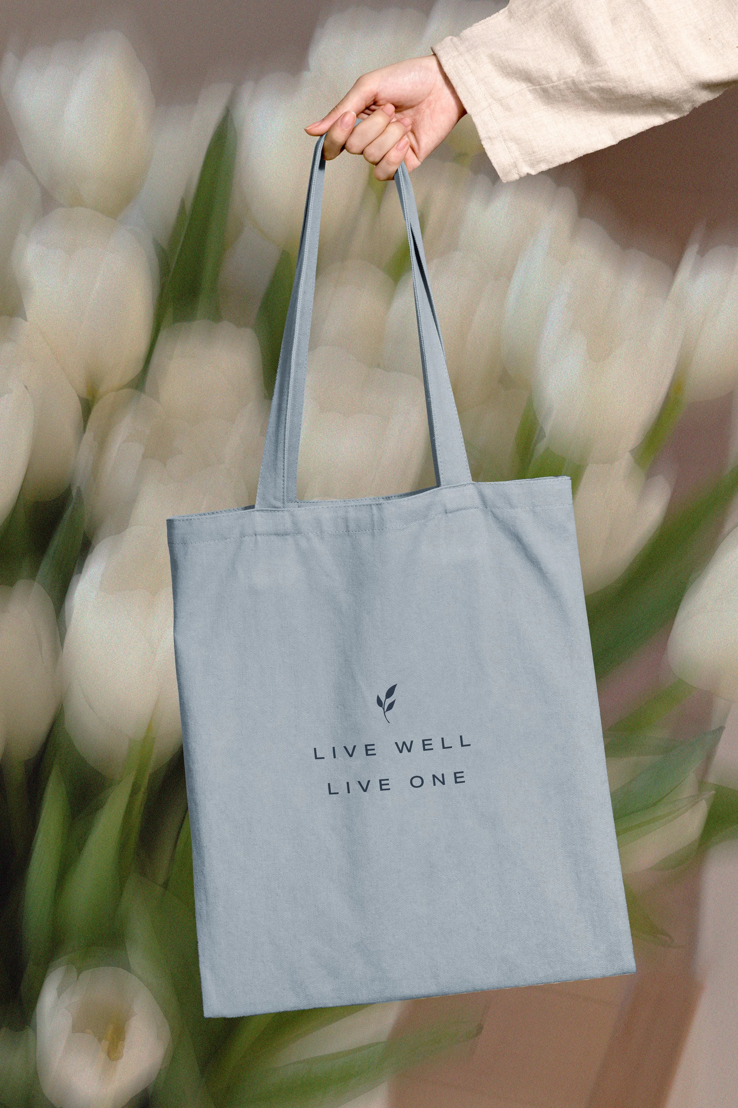 A hand holding a light gray tote bag with the text "LIVE WELL LIVE ONE" and a small leaf icon, against a background of white flowers with green stems.