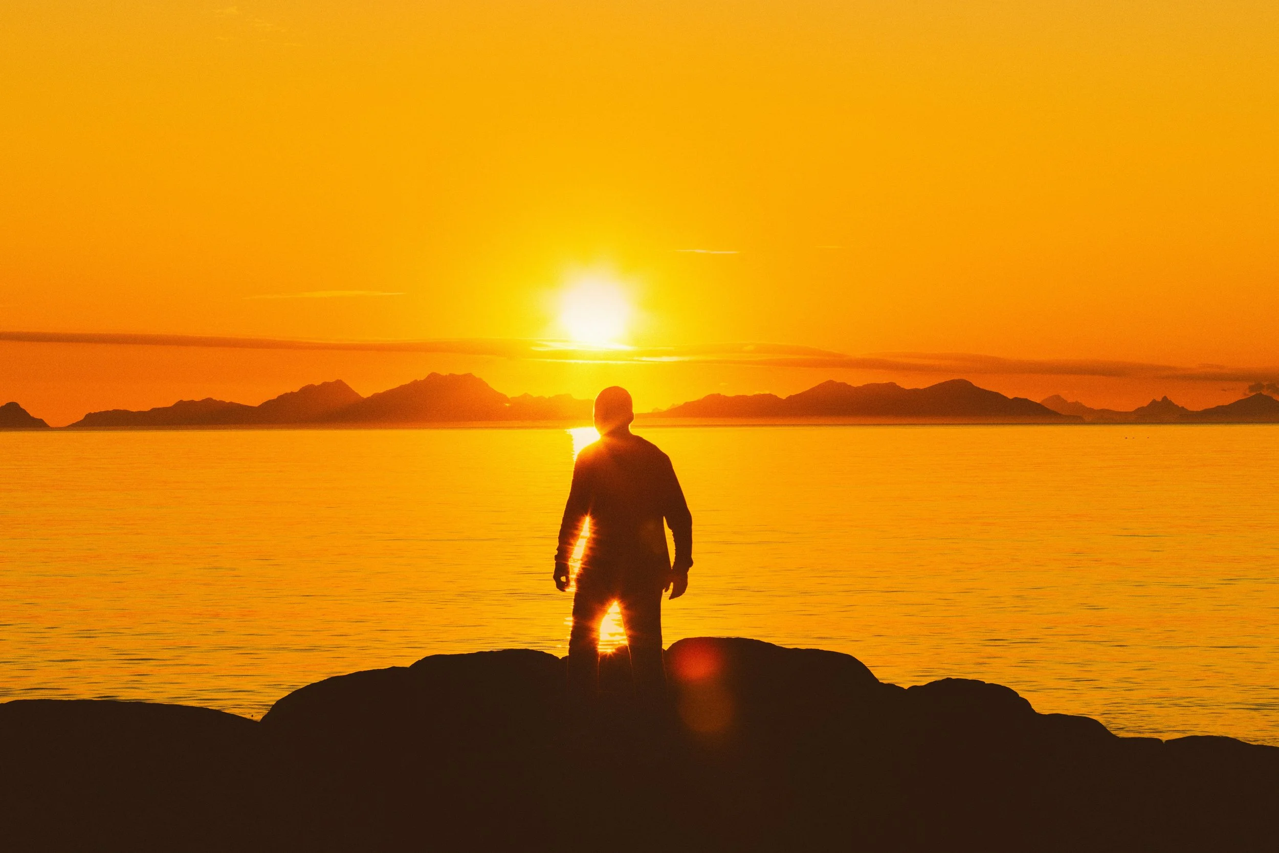 Silhouette of a man standing on rocks facing a golden sunset over the water, representing stress reduction and recovery from infrared sauna therapy