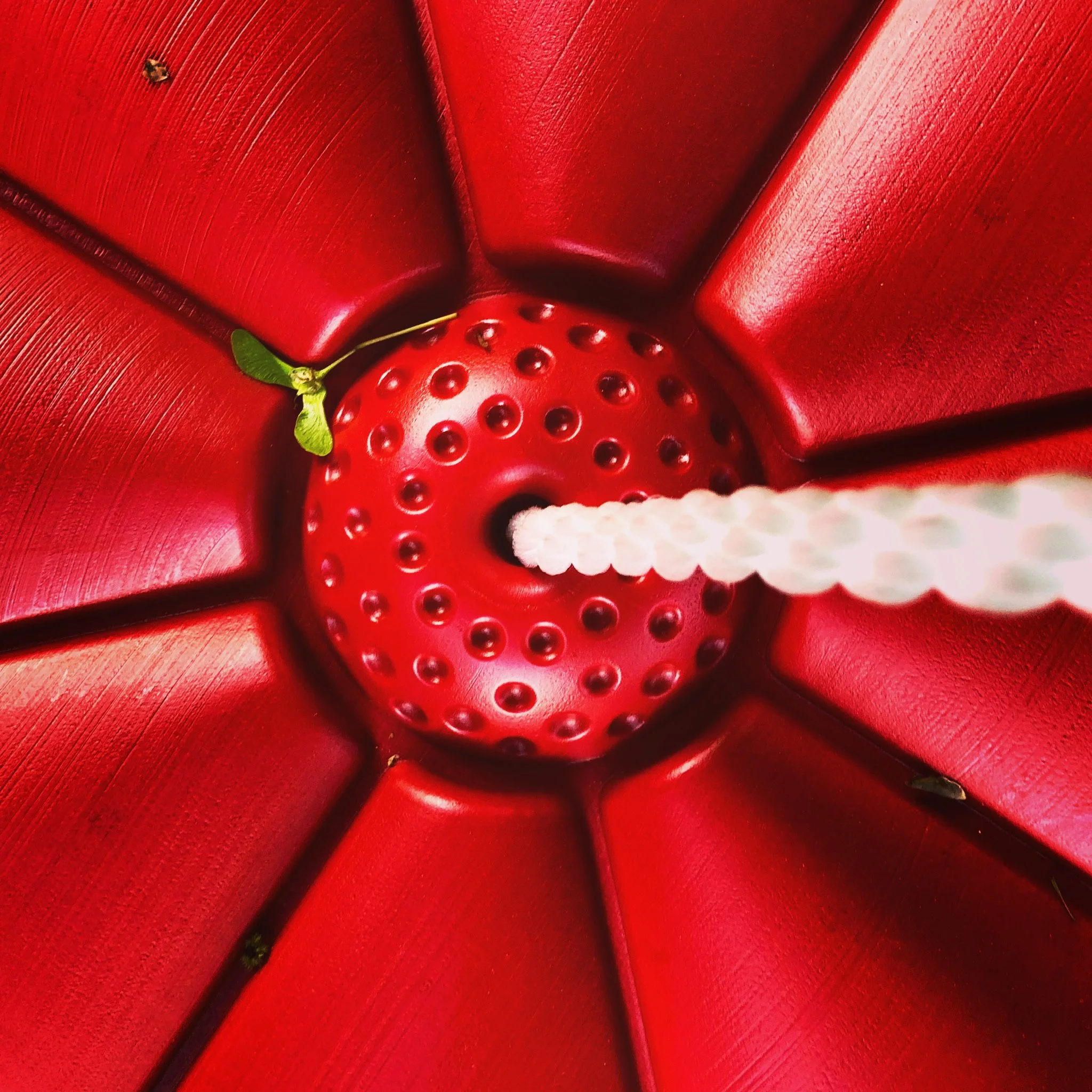Top-down view of a red patio umbrella with a white, textured straw protruding from the hole at its center. A small green leaf with a tiny insect is visible on the umbrella.