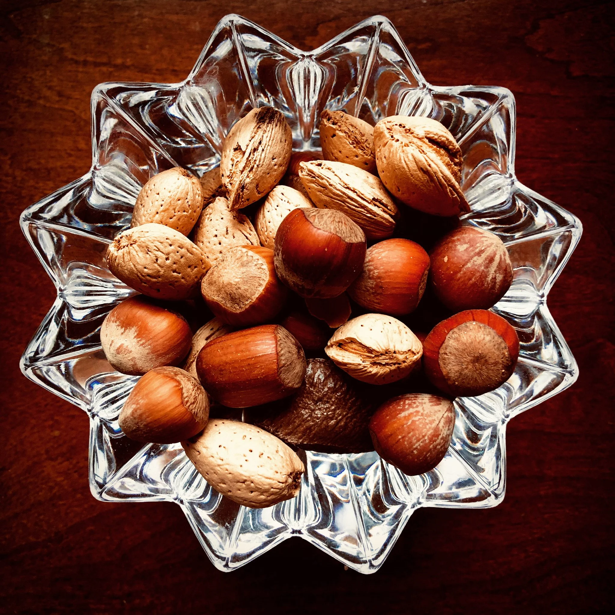 A crystal bowl filled with mixed nuts, including almonds, hazelnuts, and peanuts, on a wooden surface.